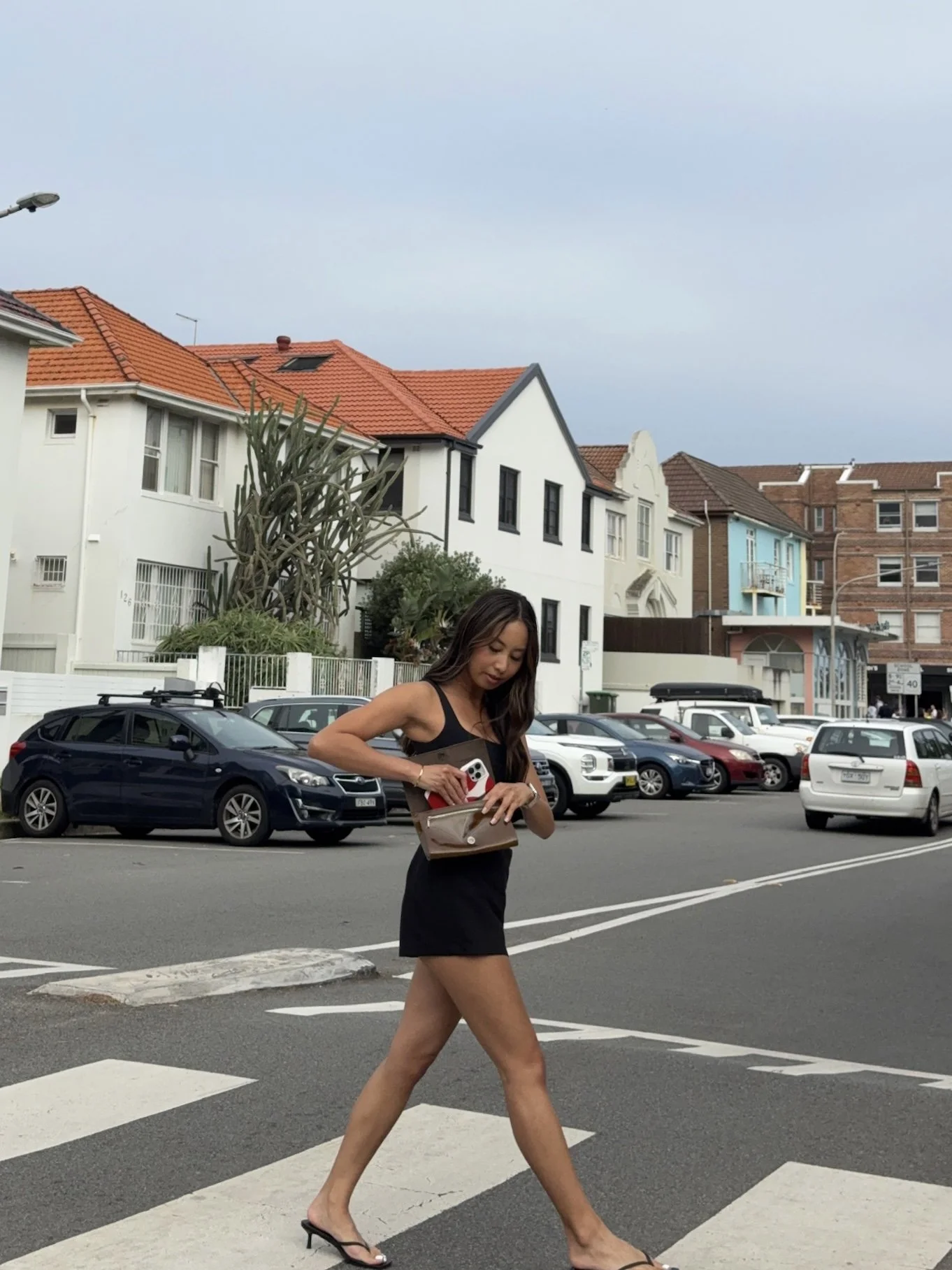 A woman in a black dress and heels crossing a street with parked cars and colorful houses in the background.