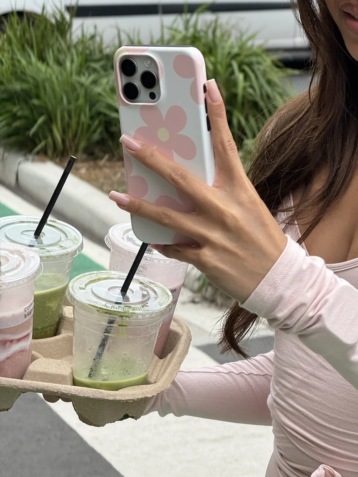 Person holding a pink and white flower-patterned phone with a tray of colorful drinks, including green, pink, and purple smoothies, outdoors.