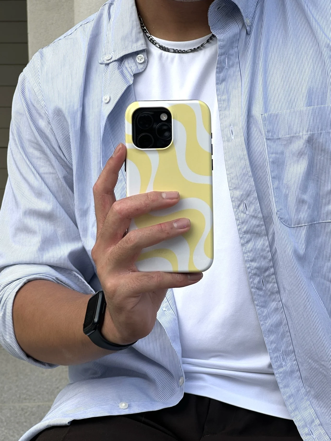 Person taking a selfie outdoors wearing a light blue striped shirt, white t-shirt, black watch, and chain necklace, holding a yellow and white patterned smartphone.