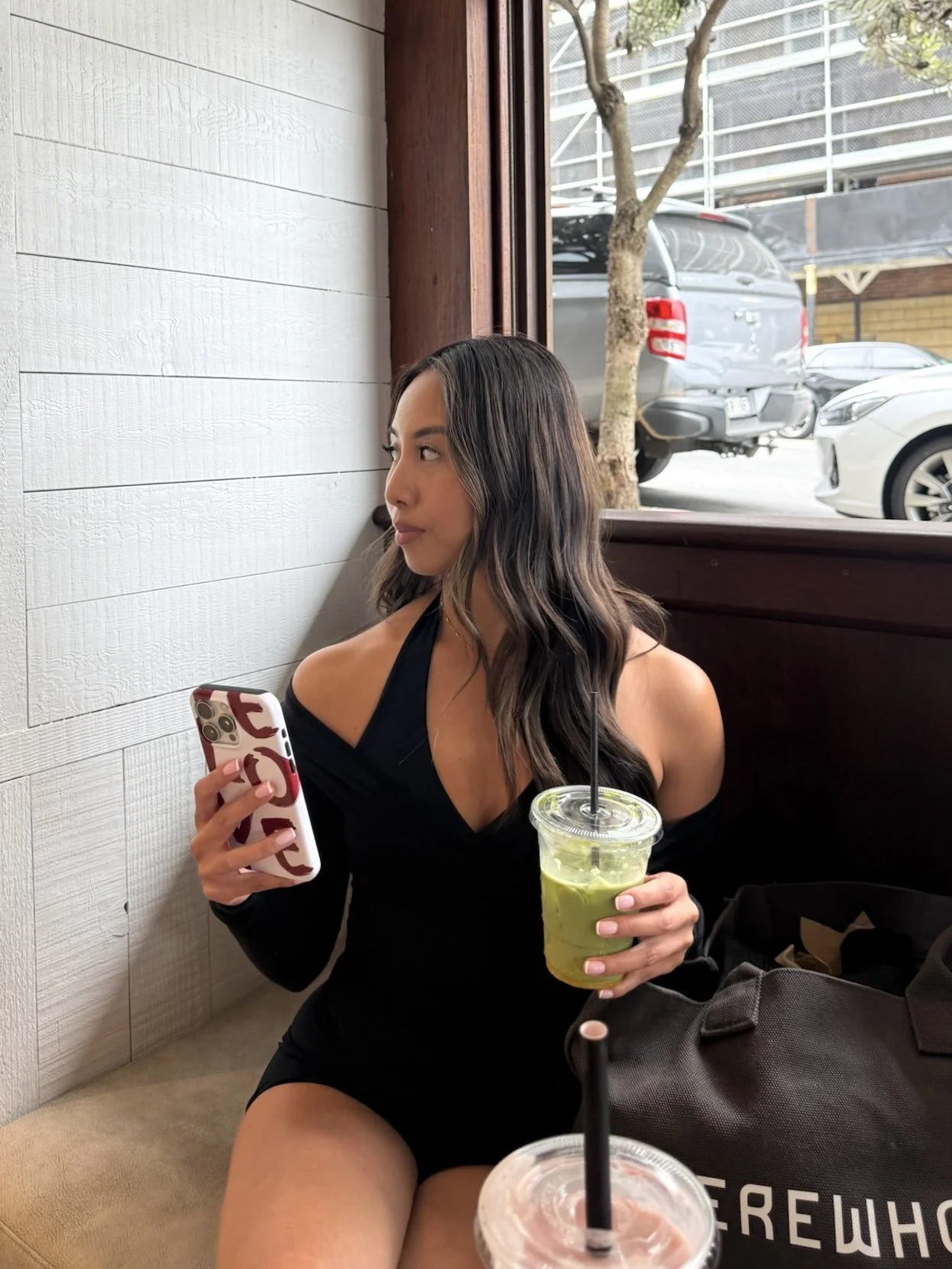 Young woman with long wavy dark hair sitting in a cafe, wearing a black off-shoulder dress, holding a green iced drink with a black straw in one hand and a smartphone with a red and white patterned case in the other, looking out the window.