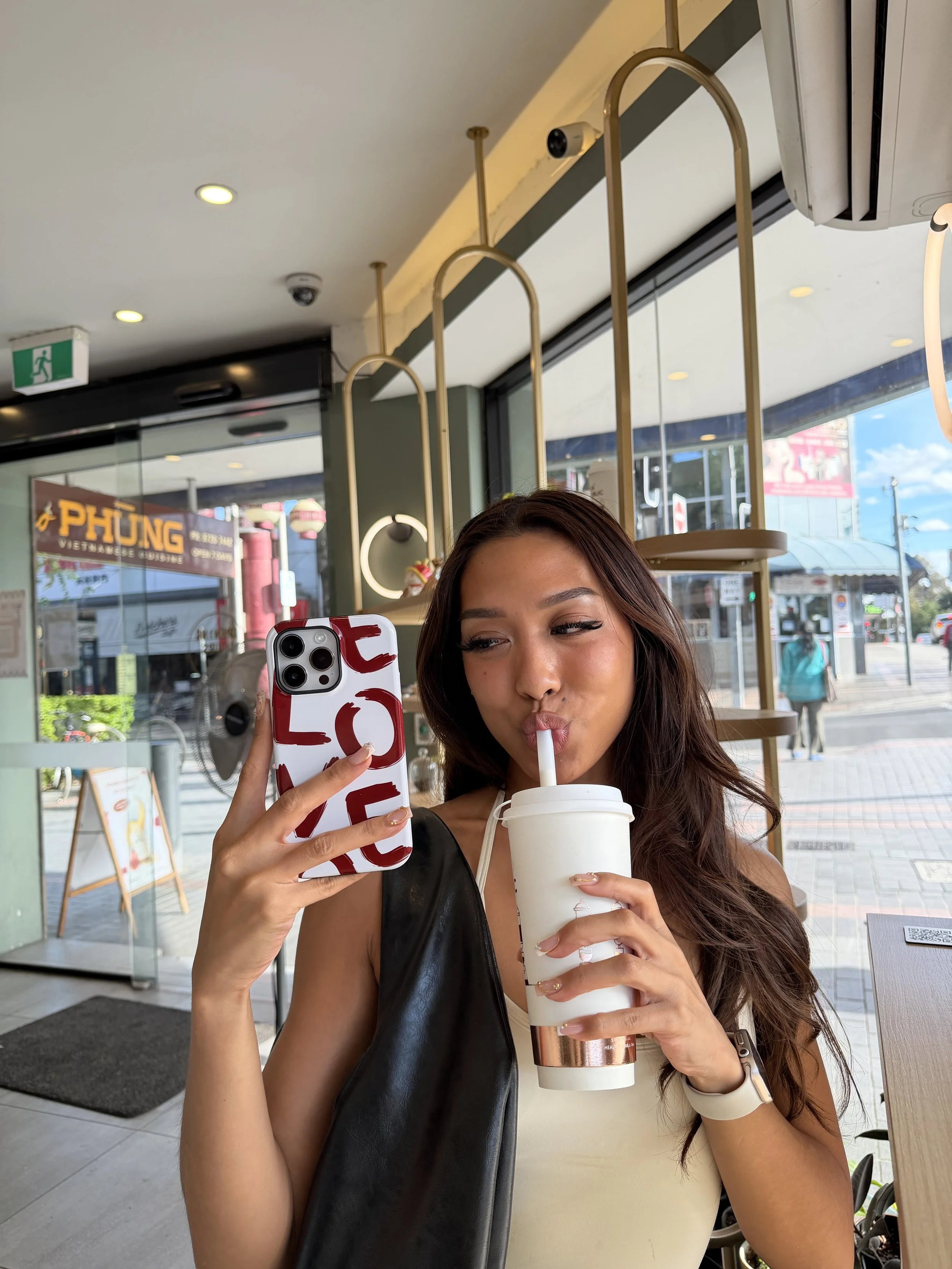 A woman sitting inside a cafe, drinking a large beverage from a cup with a straw, and taking a selfie with her smartphone.