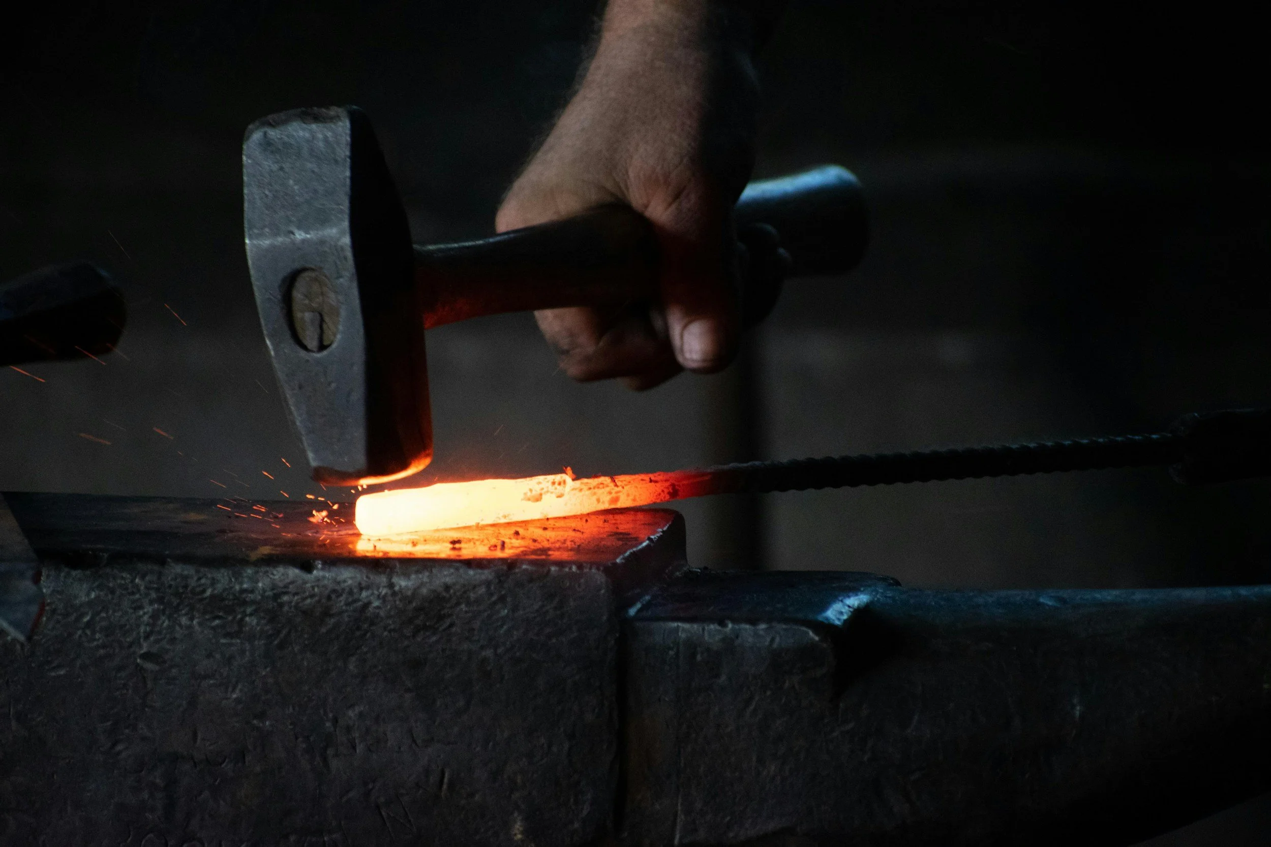 A person using a hammer to forge a heated piece of metal in a dark workshop.