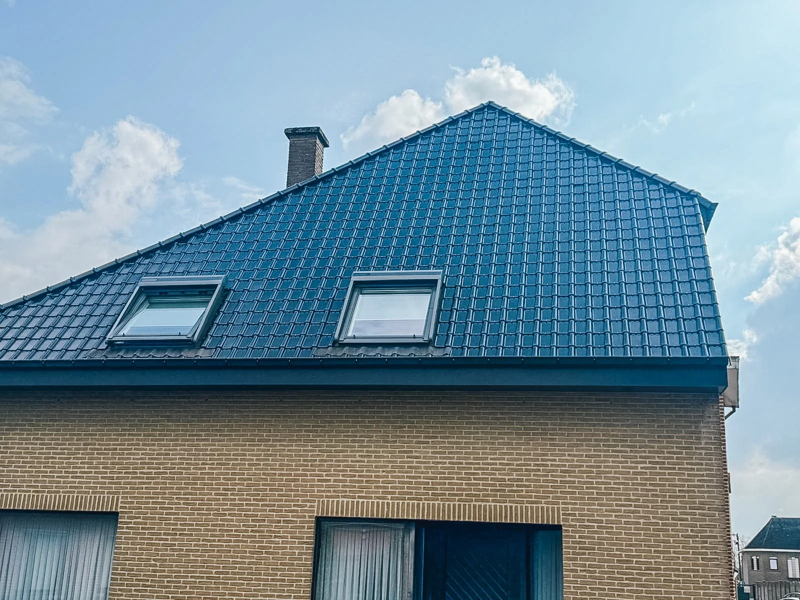 Een huis met een bruine bakstenen muur en een blauw metalen zadeldak met twee dakramen, onder een heldere blauwe hemel met enkele witte wolken.