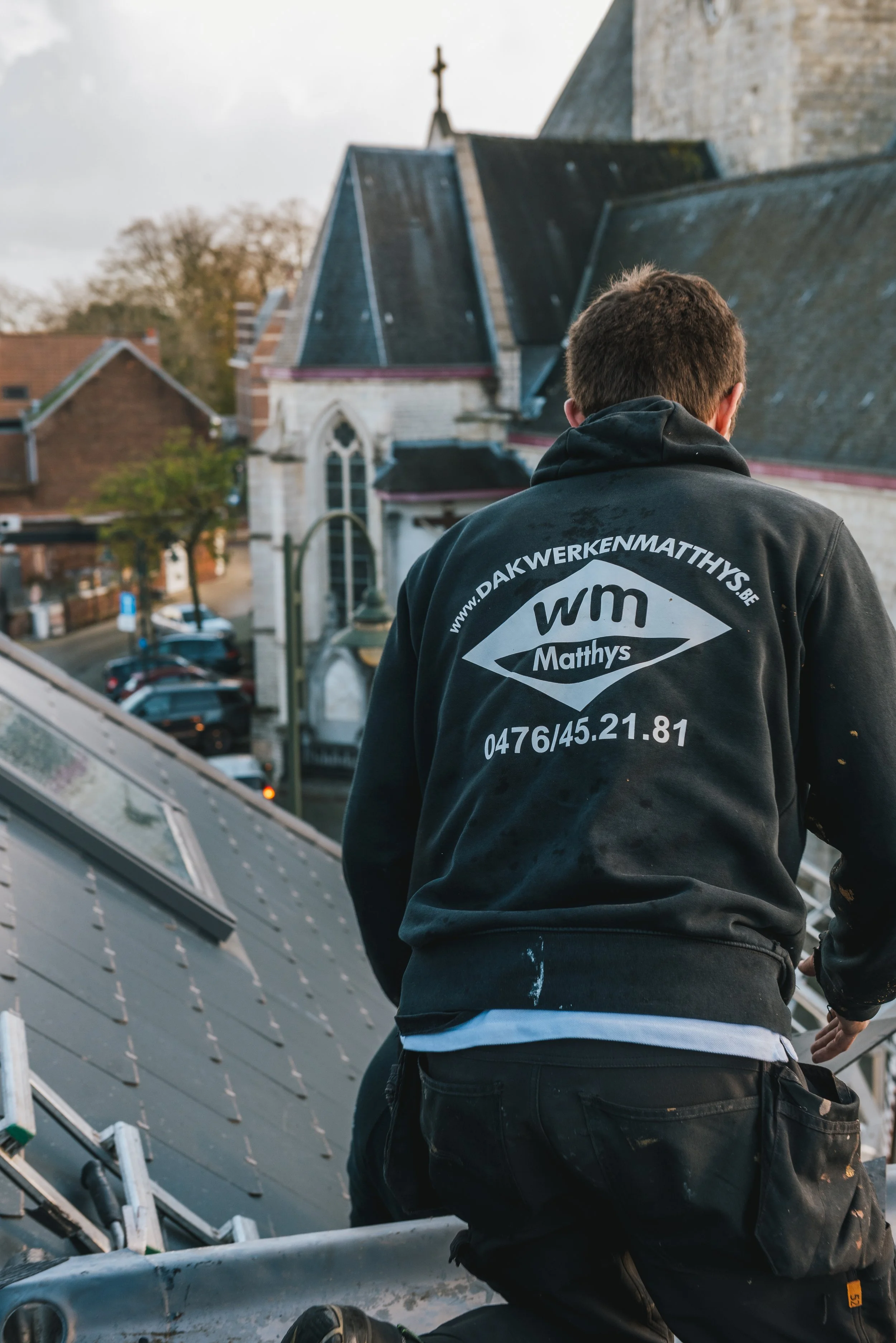 Een man die op het dak van een gebouw werkt, gekleed in een zwart shirt met de tekst en contactgegevens van een dakbedekkingsbedrijf.