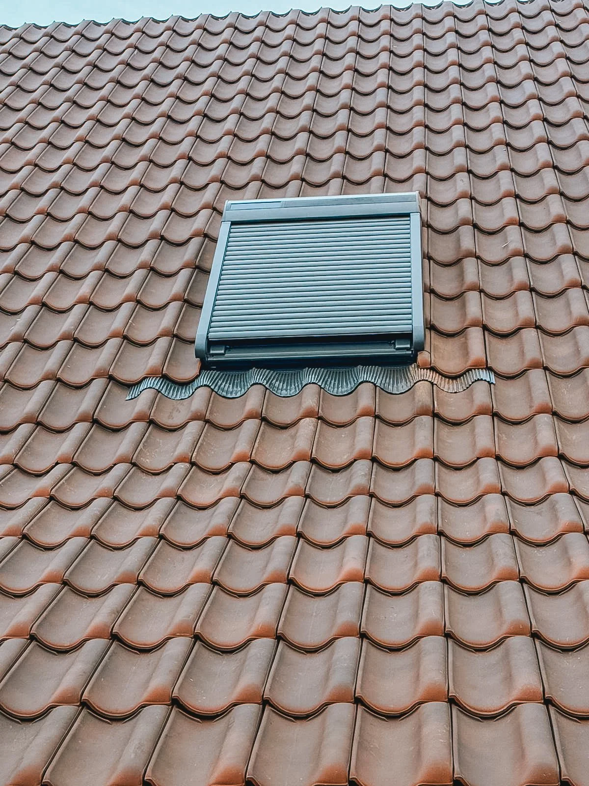 Dak met keramische terracotta dakpannen en een plat dakraampje met een rolluik.