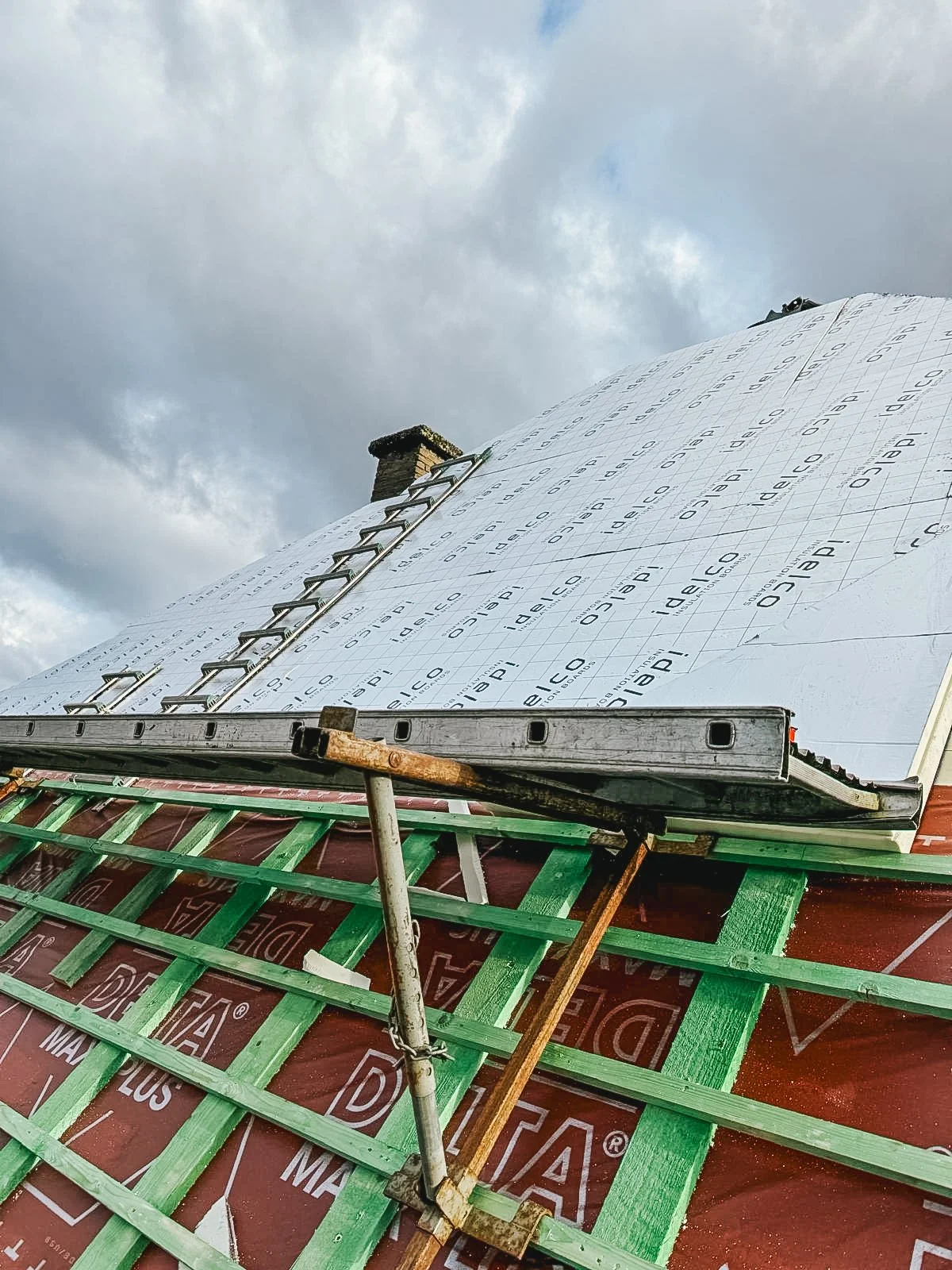 Schildering van een dak onder constructie, met een ladder en bouwmaterialen, tegen een bewolkte hemel