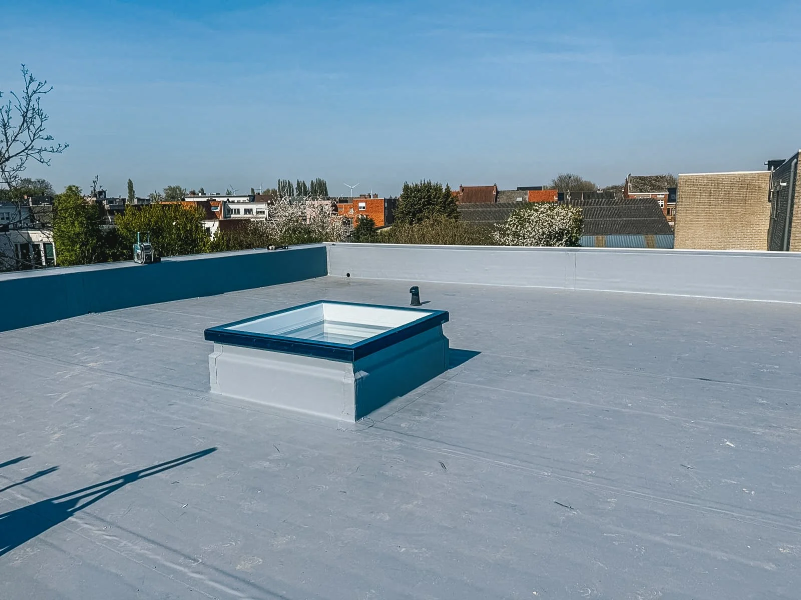 Uitzicht vanaf een platte, gepantserde dakterras met een dakraam, gericht op een rustige woonwijk met huizen en bomen onder een heldere blauwe hemel.