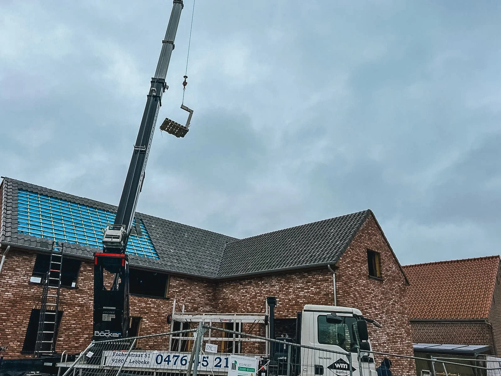 Bouwkraan die een pallet optilt naast een rood bakstenen huis met zonnepanelen op het dak.