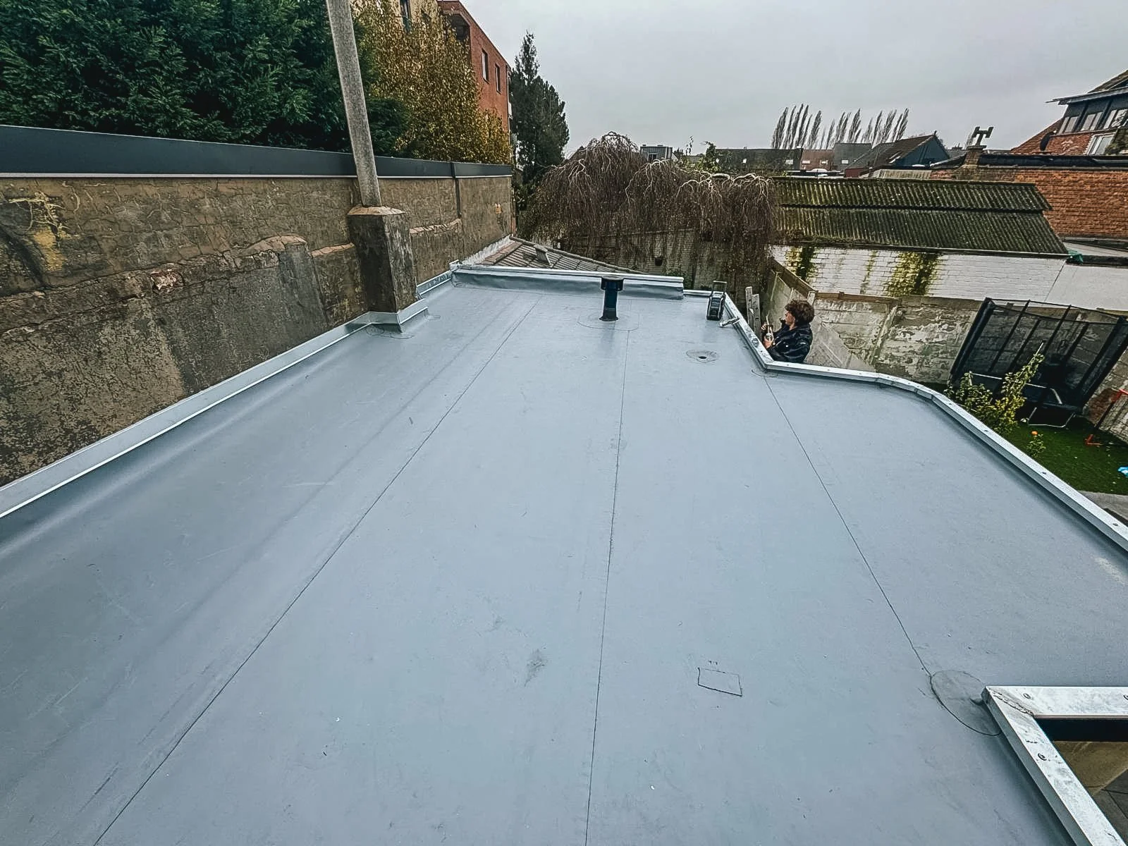 Op het platte dak van een gebouw wordt een nieuwe waterdichte coating aangebracht. Een man staat op de achtergrond, die met een gereedschap bezig is.