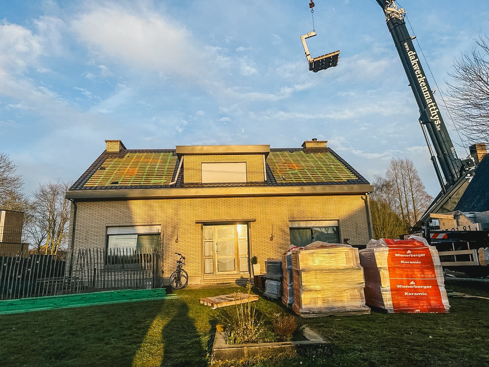 Een huis onder renovatie met nieuwe dakpannen en bouwmaterialen voor dakwerk. Er staat een bouwkraan op het terrein.