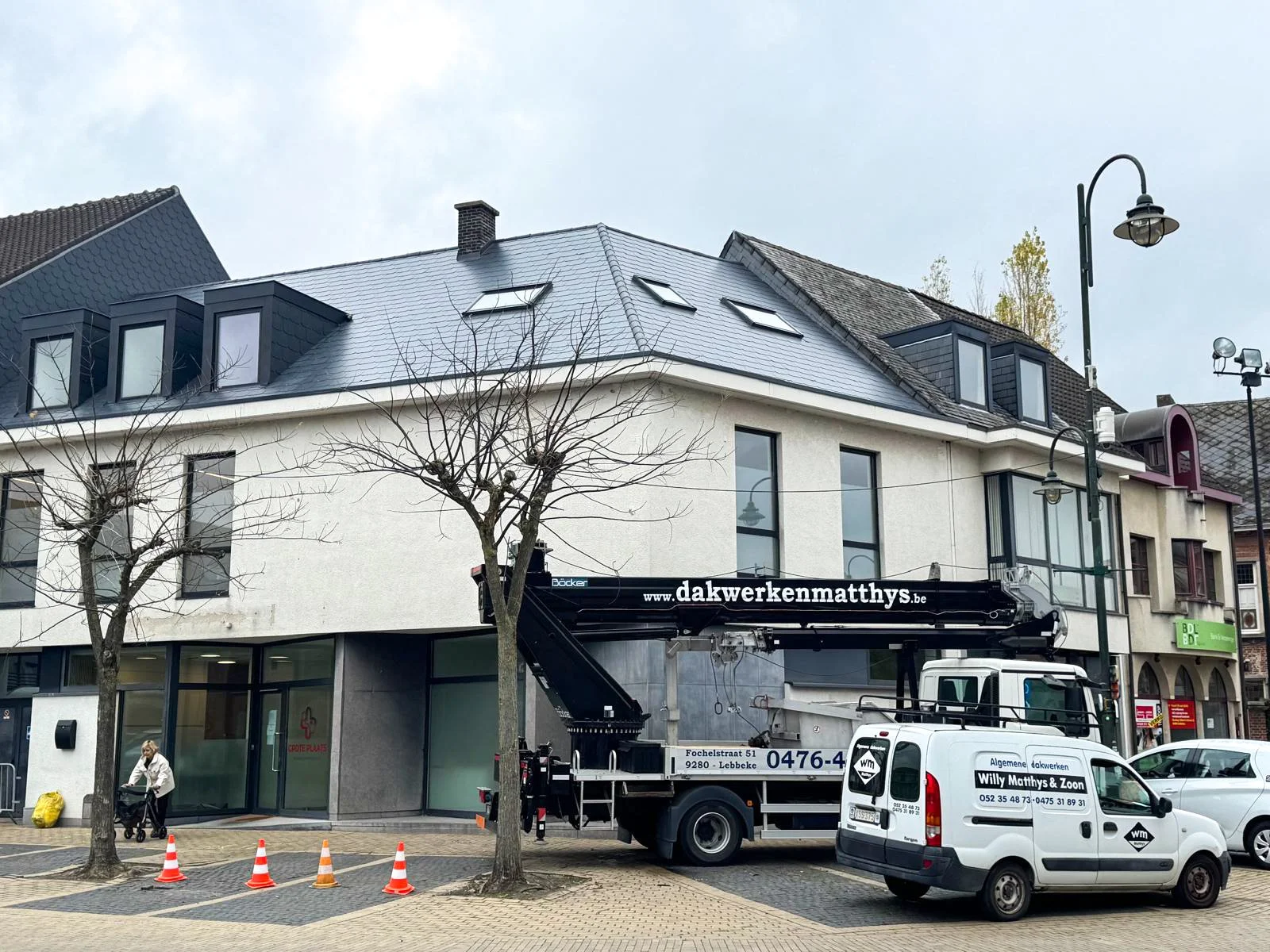 Een wit gebouw met grote ramen en een zwarte dakgoot, twee kale bomen op de stoep, een vrouw met een kinderwagen, en een grote kraanwagen van een dakdekkersbedrijf voor de gevel. 