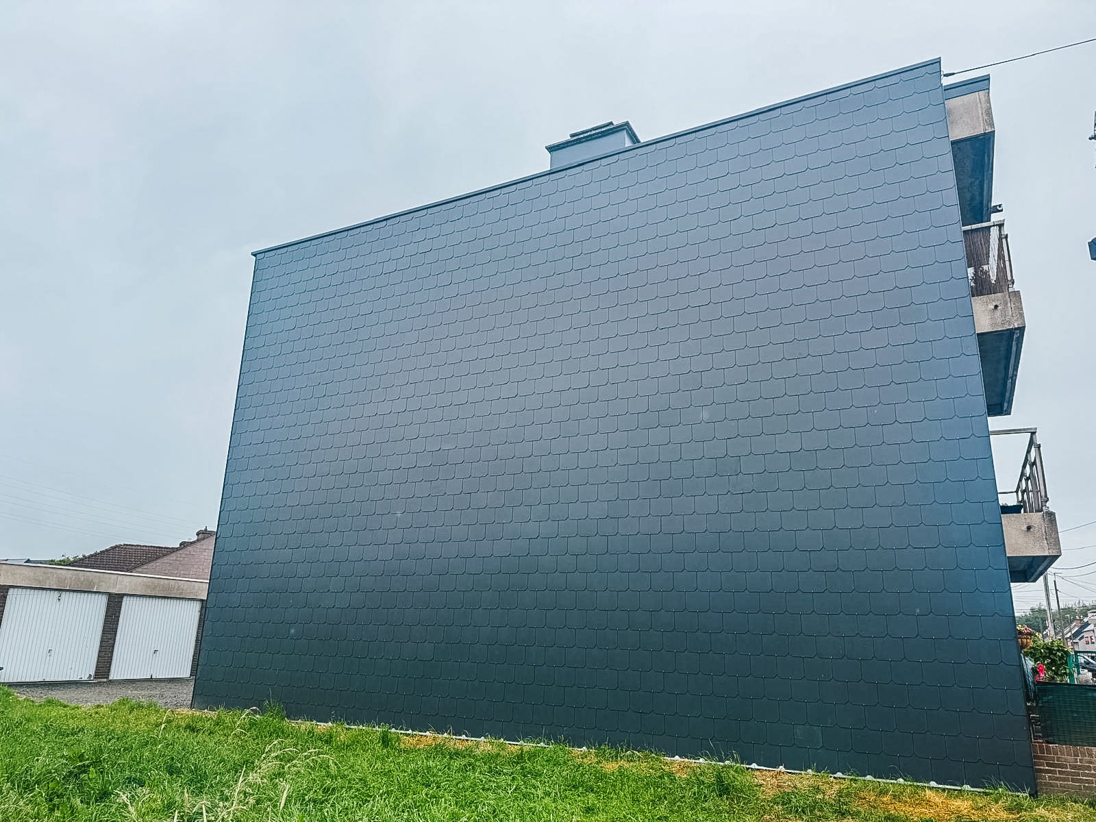 Een groot, modern gebouw met een gevel bedekt met zwarte dakpanvormige tegels, een klein stukje gras voor het gebouw en een kleine witte omheining links.
