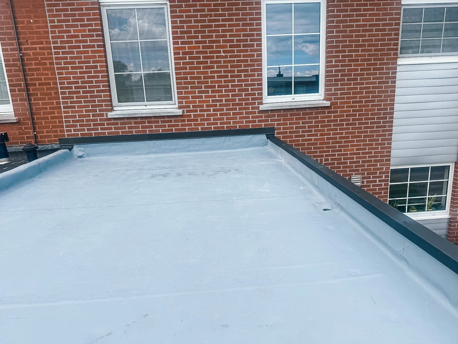 Opgenomen van een plat dak met een lichte coating, vooraan een zwarte afwerking, en een rode bakstenen gebouw met vier ramen in de achtergrond.