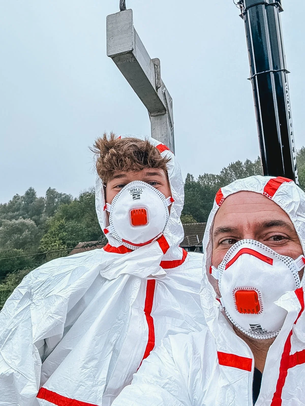 Twee mensen in volle persoonlijke beschermingsuitrusting, inclusief witte overalls en ademhalingsmaskers, staan buiten op een bewolkte dag, met een heuvel en bomen op de achtergrond.
