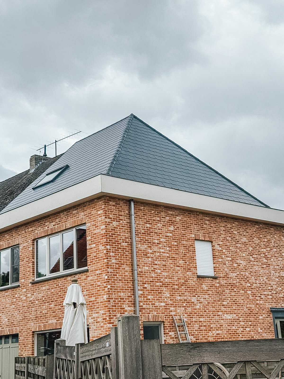 A brick house with a tiled sloped roof, a skylight, and a window with a closed white shutter. There is a ladder leaning against the wall and a patio umbrella outside.