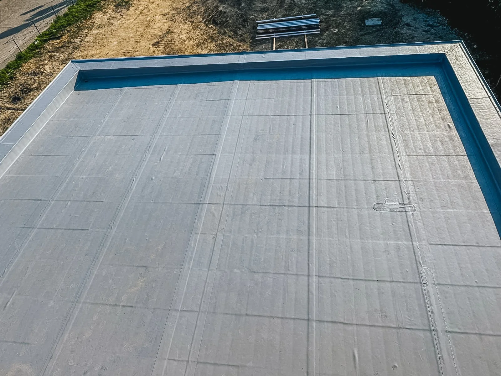 Uitzicht van een uit de lucht gefotografeerd plat dak met witte waterdichte laag, omringd door een rand met een blauwe coating, en een stuk aangelegd terrein met wat gras en een weg ernaast.