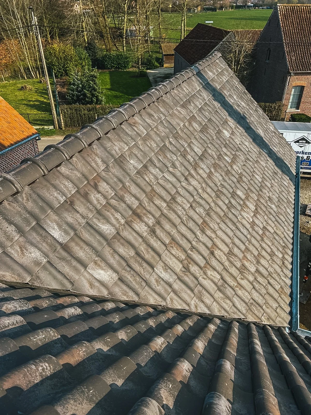 Uitzicht op een dak met betonnen dakpannen, omgeven door andere daken en een tuin met gras en bomen.