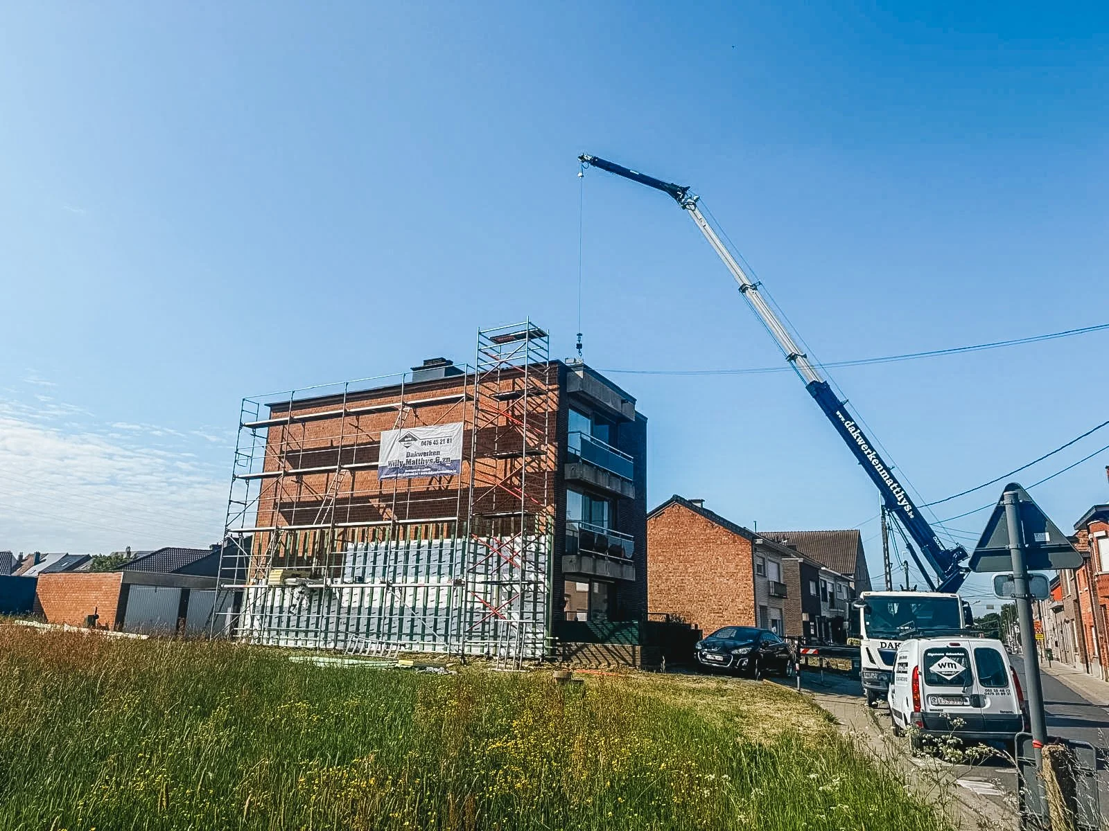 Een bouwplaats met een hoogbouwproject in aanbouw, een grote kraan die materialen omhoog tilt, een steiger rondom het gebouw en een paar geparkeerde auto's, onder een heldere blauwe hemel.