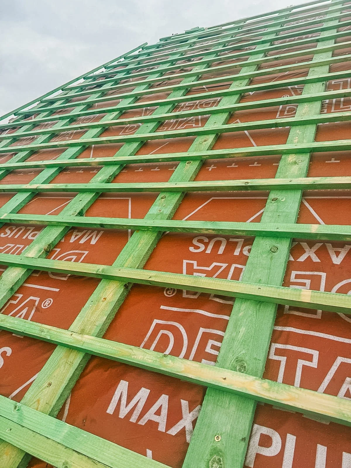 Constructie van een dak met houten latten en isolatiemateriaal eronder, onder een grijze wolkenlucht.