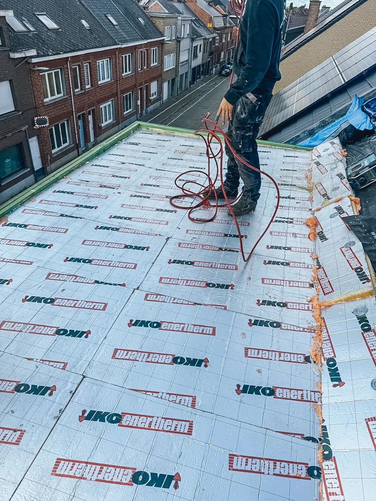 Persoon op een dak die isolatiemateriaal aanbrengt met kabels. In de achtergrond is een straat met huizen te zien.