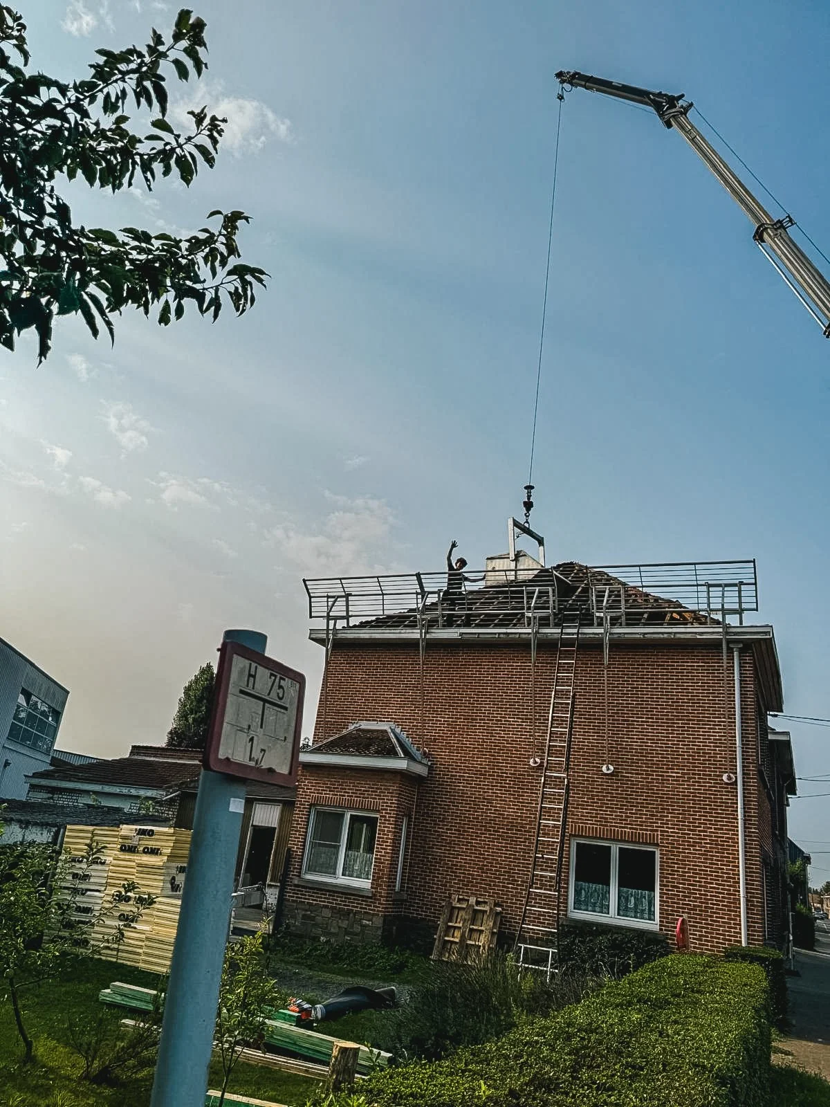 Een huis in aanbouw met een hijskraan die een constructie boven op het dak tilgt; er staat een persoon op het dak die zwaaien naar de camera; er is een temperature gauge zichtbaar in de voorgrond.