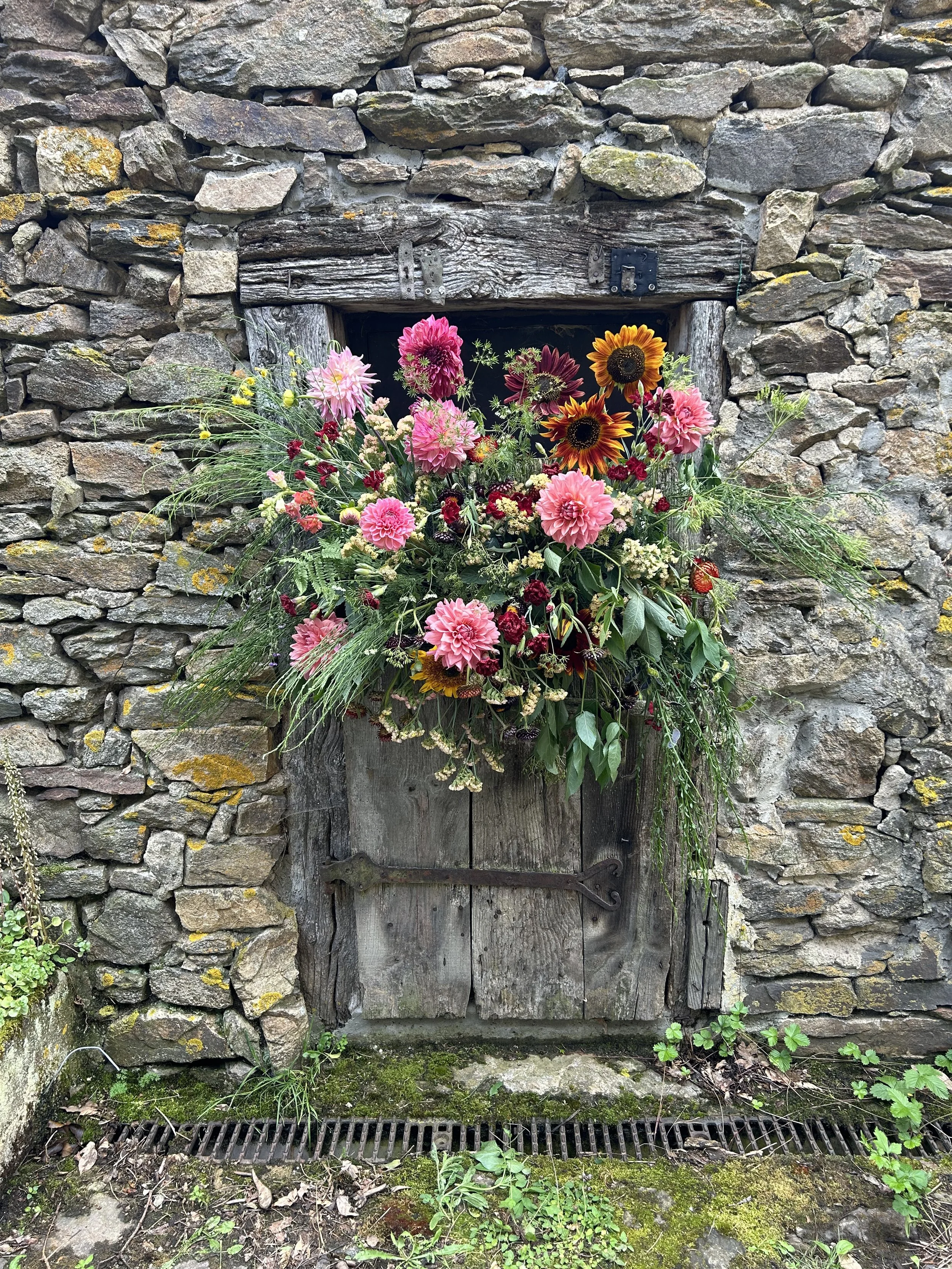 Une vieille porte en bois avec une grande composition florale colorée devant. La porte est encastrée dans un mur en pierre.