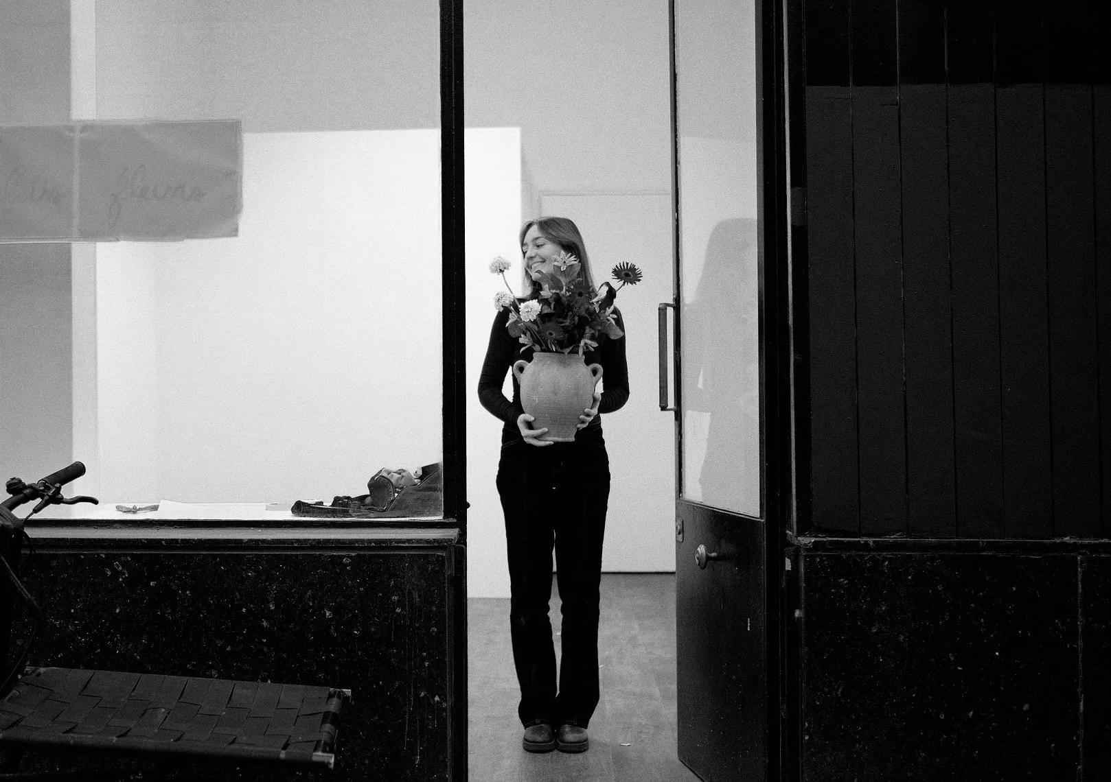 Une femme souriante tient un vase avec des fleurs, vue à travers une vitre depuis une pièce sombre, en noir et blanc.