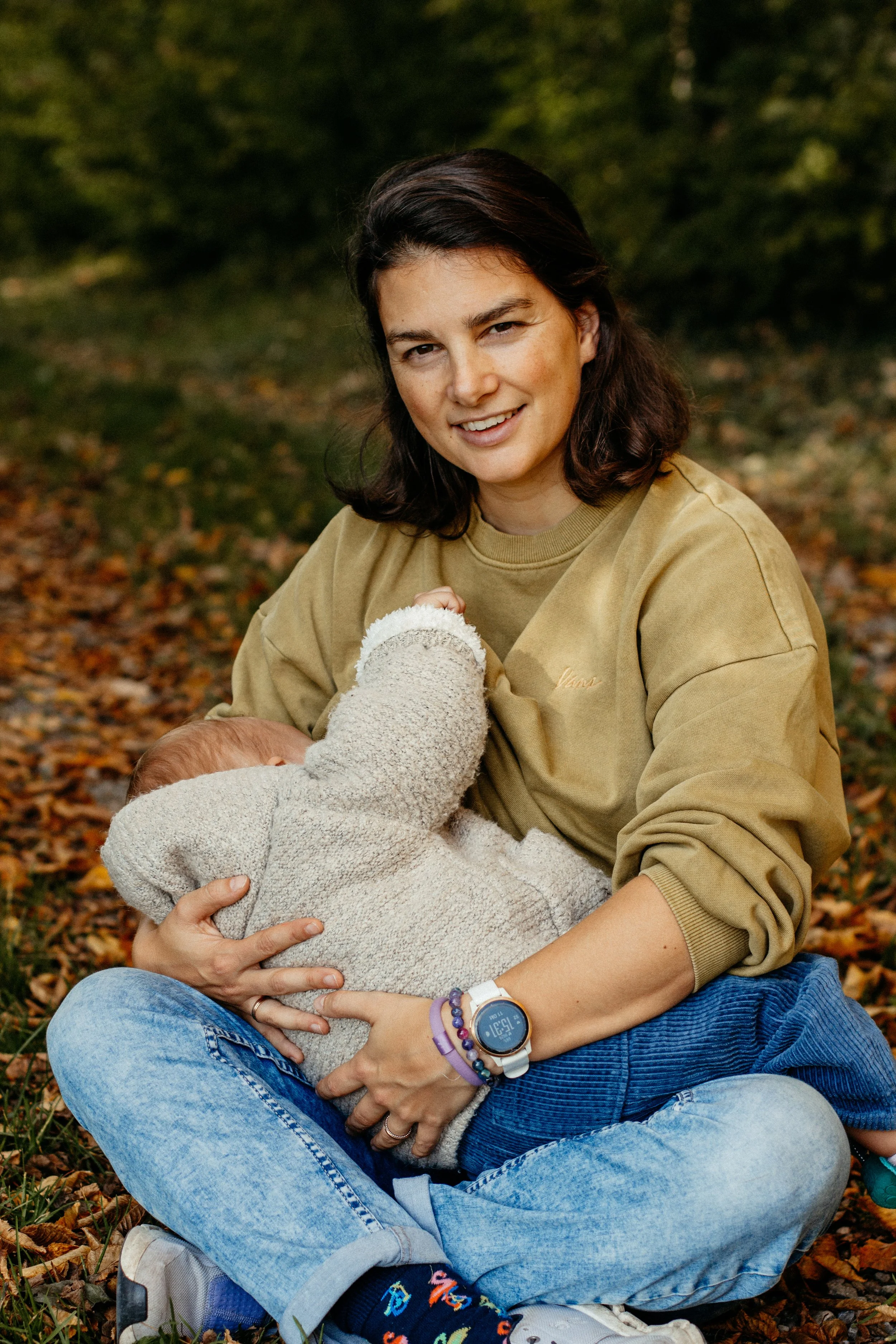 Eine Frau sitzt im Freien, hält ein Baby im Arm, umgeben von herbstlichen Blättern, lächelt in die Kamera.