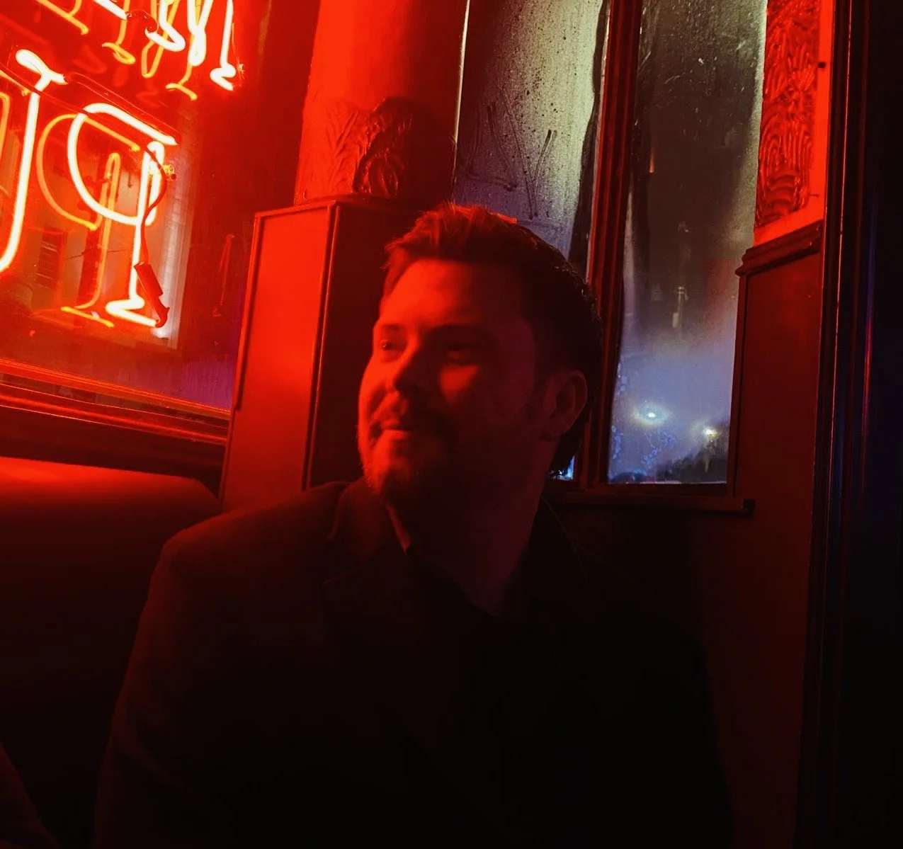 A man sitting in a dimly lit space illuminated by red neon lights, with a neon sign reflecting on the window behind him.