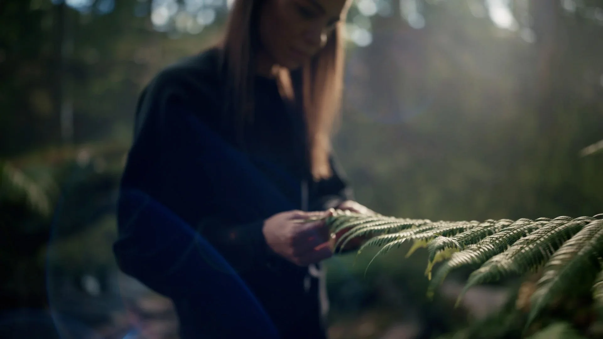 Eine Frau berührt grüne Blätter in einem Wald bei Sonnenlicht.