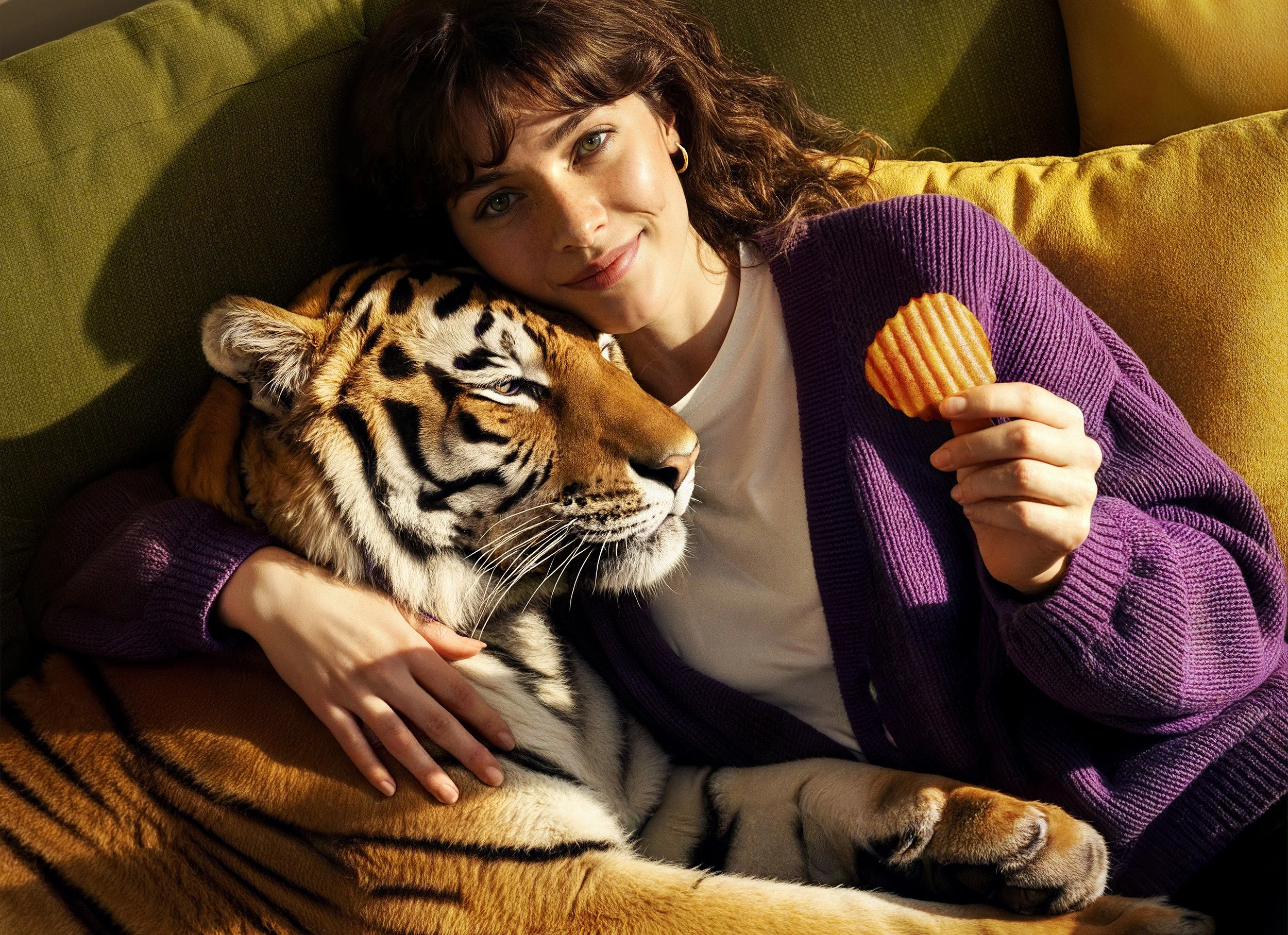 Junge Frau mit braunen, lockigen Haaren, die auf einer grünen Couch mit einem kleinen Jaguar zu sehen ist, während sie ein Kartoffelchip in der Hand hält. Das Licht fällt auf sie und den Panther.