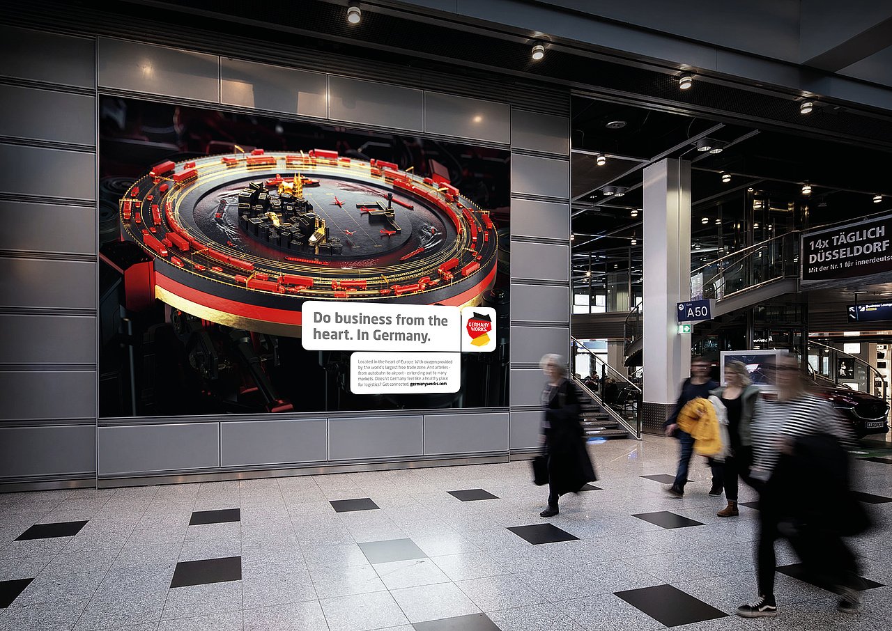Großes digitales Werbeplakat im Flughafen mit einem roten und schwarzen Industrie-Miniaturmodell und der Aufschrift 'Do business from the heart. In Germany.'