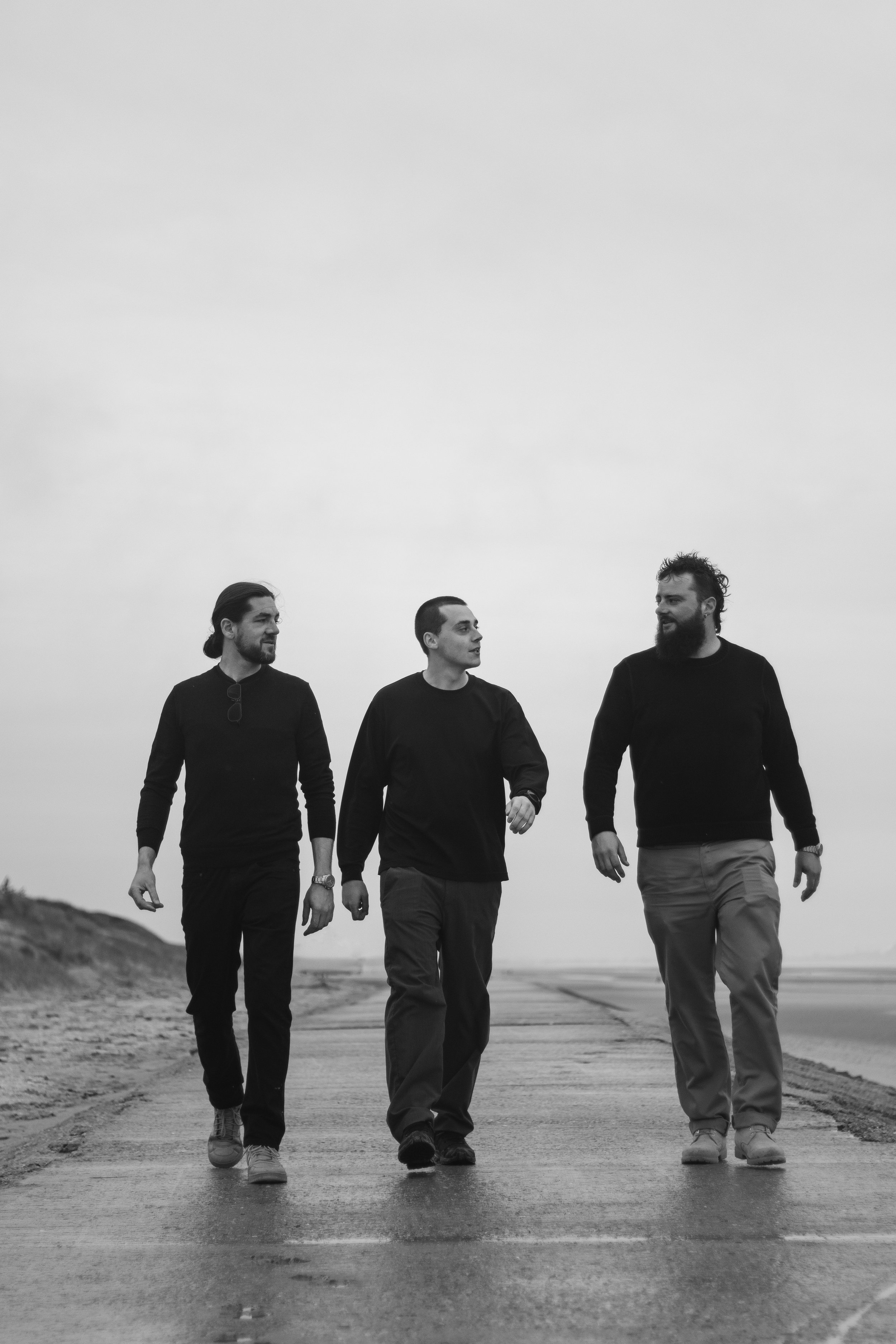 Drie mannen wandelen samen op een weg aan het strand, allemaal gekleed in donkere truien en lichte broeken, in zwart-wit.