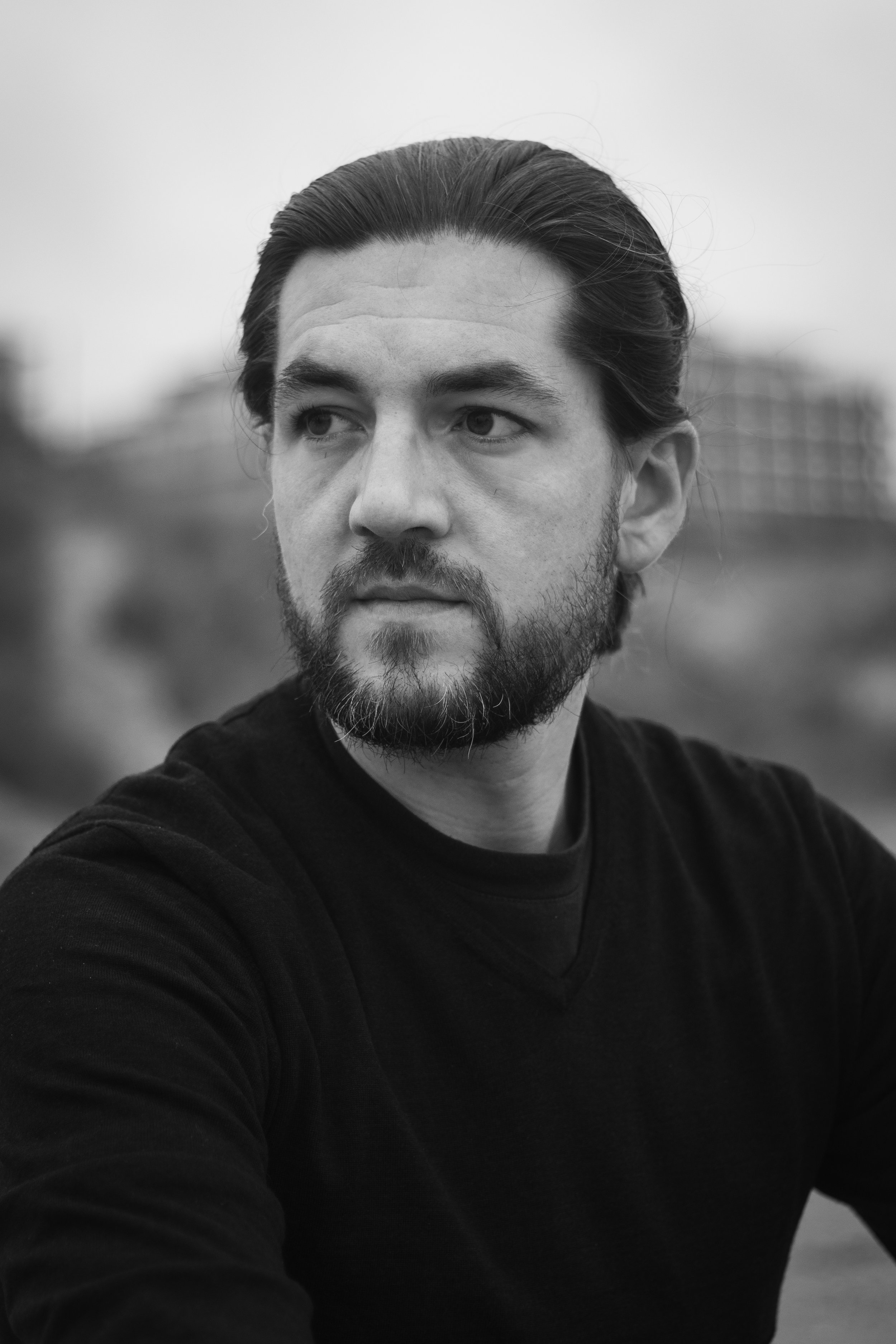Zwarte en witte portretfoto van een man met lange haar en een baard, gekleed in een zwarte trui, met een achtergrond van een gebouw en een landschap, buiten.