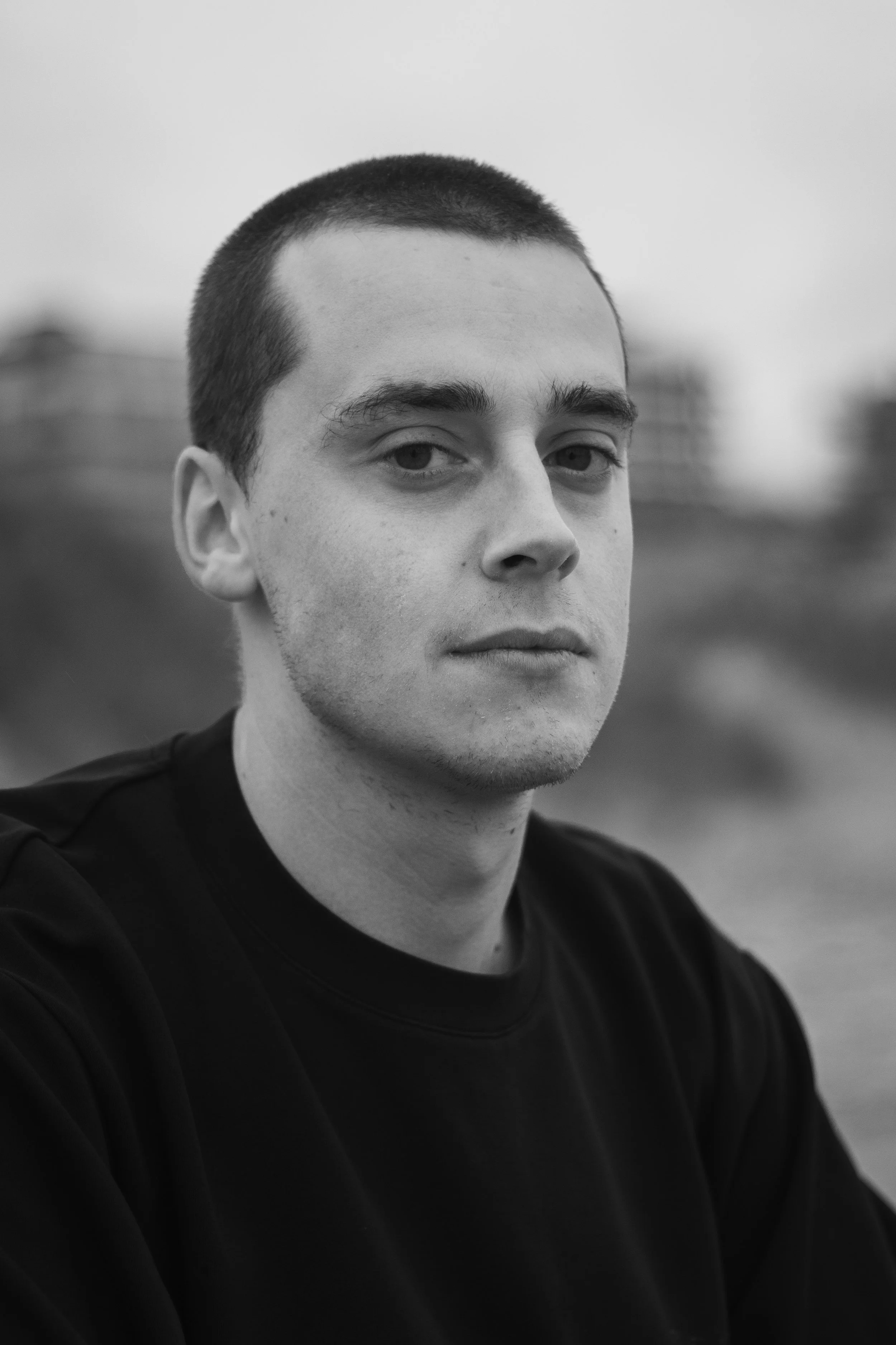 Een zwart-witportret van een jonge man met korte, donkere haar en een serieuze blik, zittend buiten.