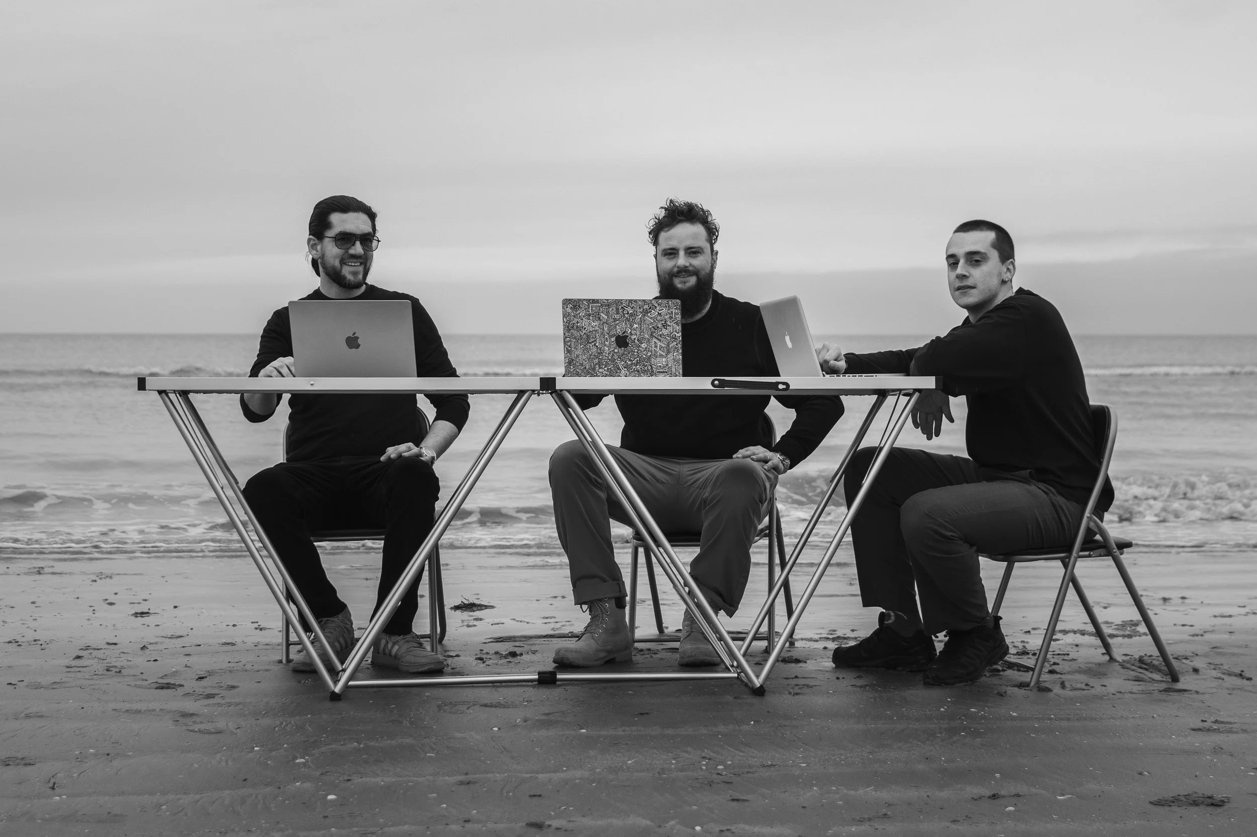 Drie mannen zitten op stoelen bij een tafel op het strand, werken met laptops, en kijken naar de camera, zee op achtergrond.