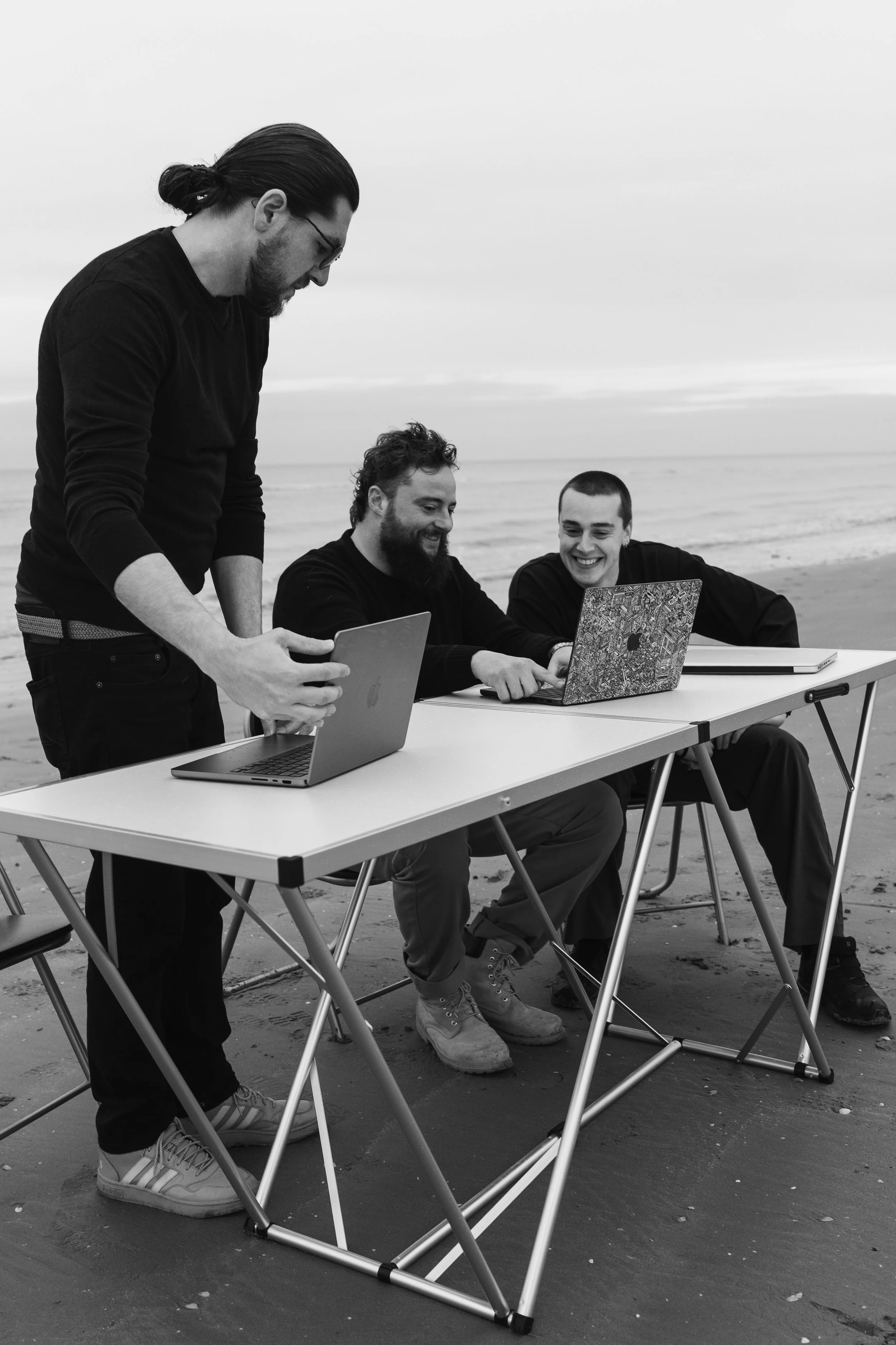 Drie jonge mannen werken samen op laptops aan een tafel op het strand, terwijl ze lachen en praten.