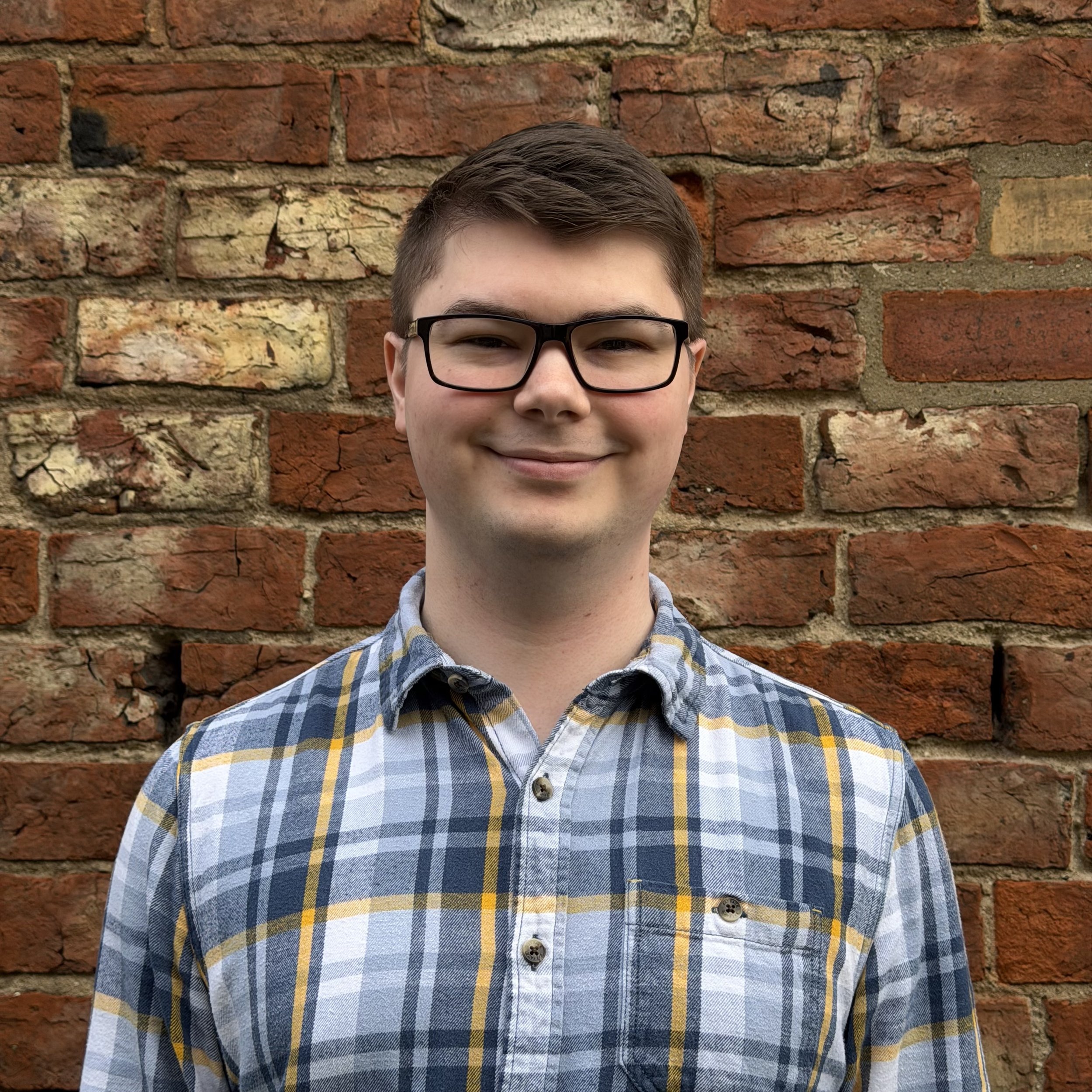 A smiling young man with short brown hair, glasses, and a plaid shirt standing in front of a red brick wall.