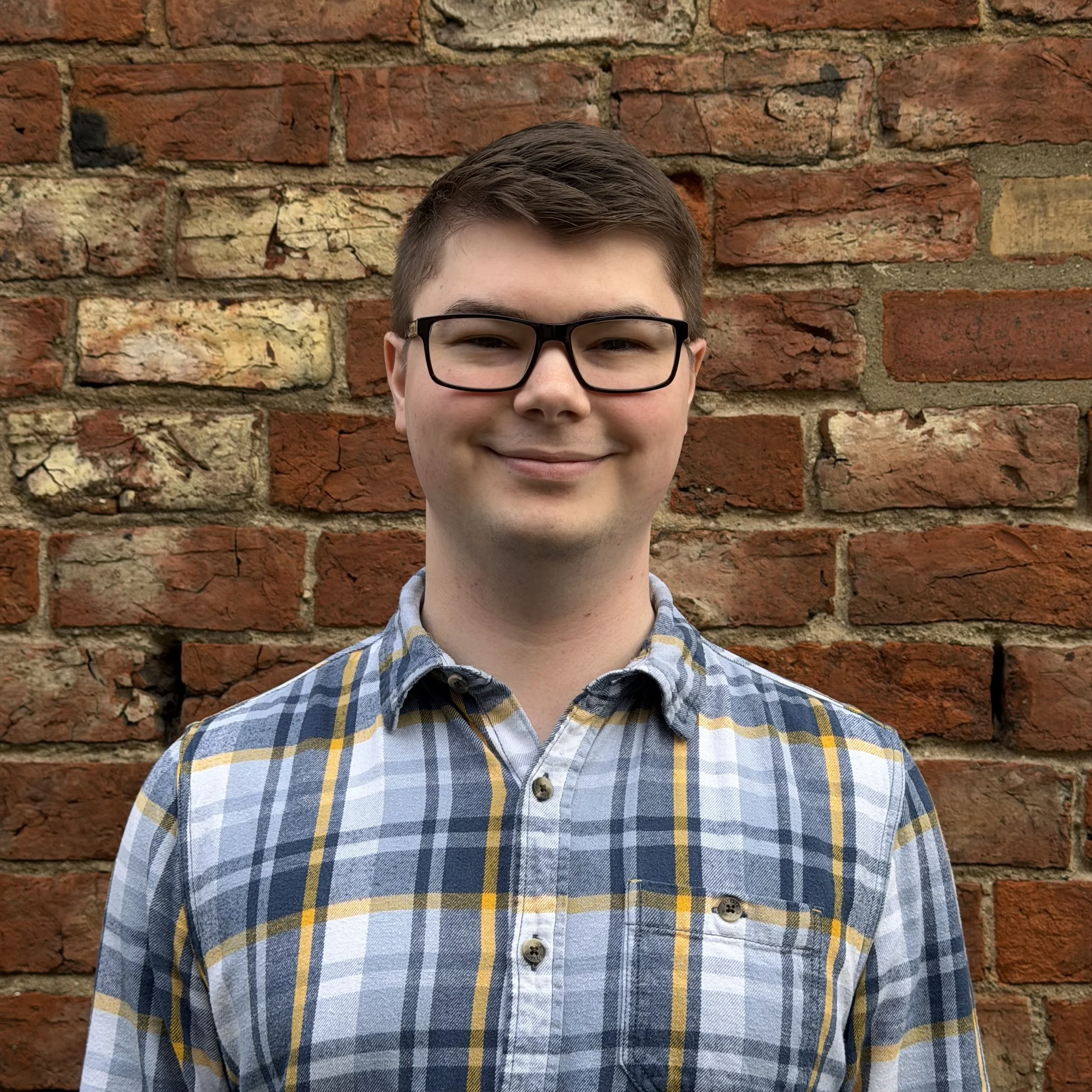 A smiling young man with short brown hair, glasses, and a plaid shirt standing in front of a red brick wall.