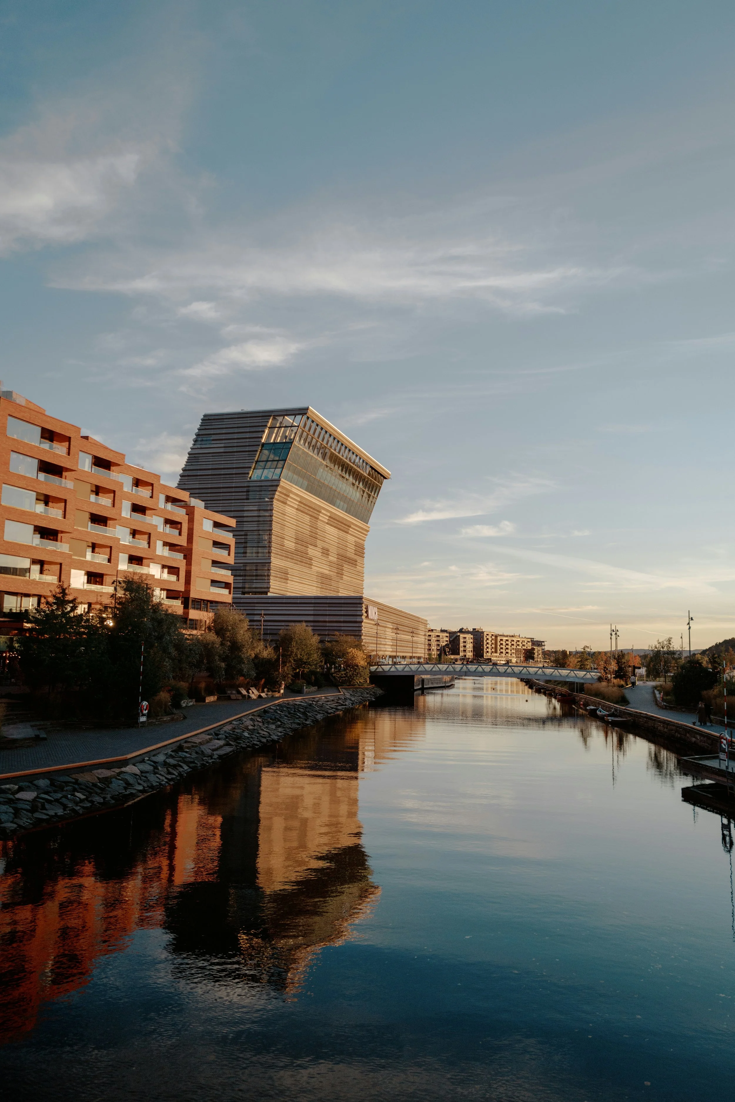 Modern cityscape with a distinctive tilted building reflected in a river, with a pedestrian walkway along the water, under a partly cloudy sky.