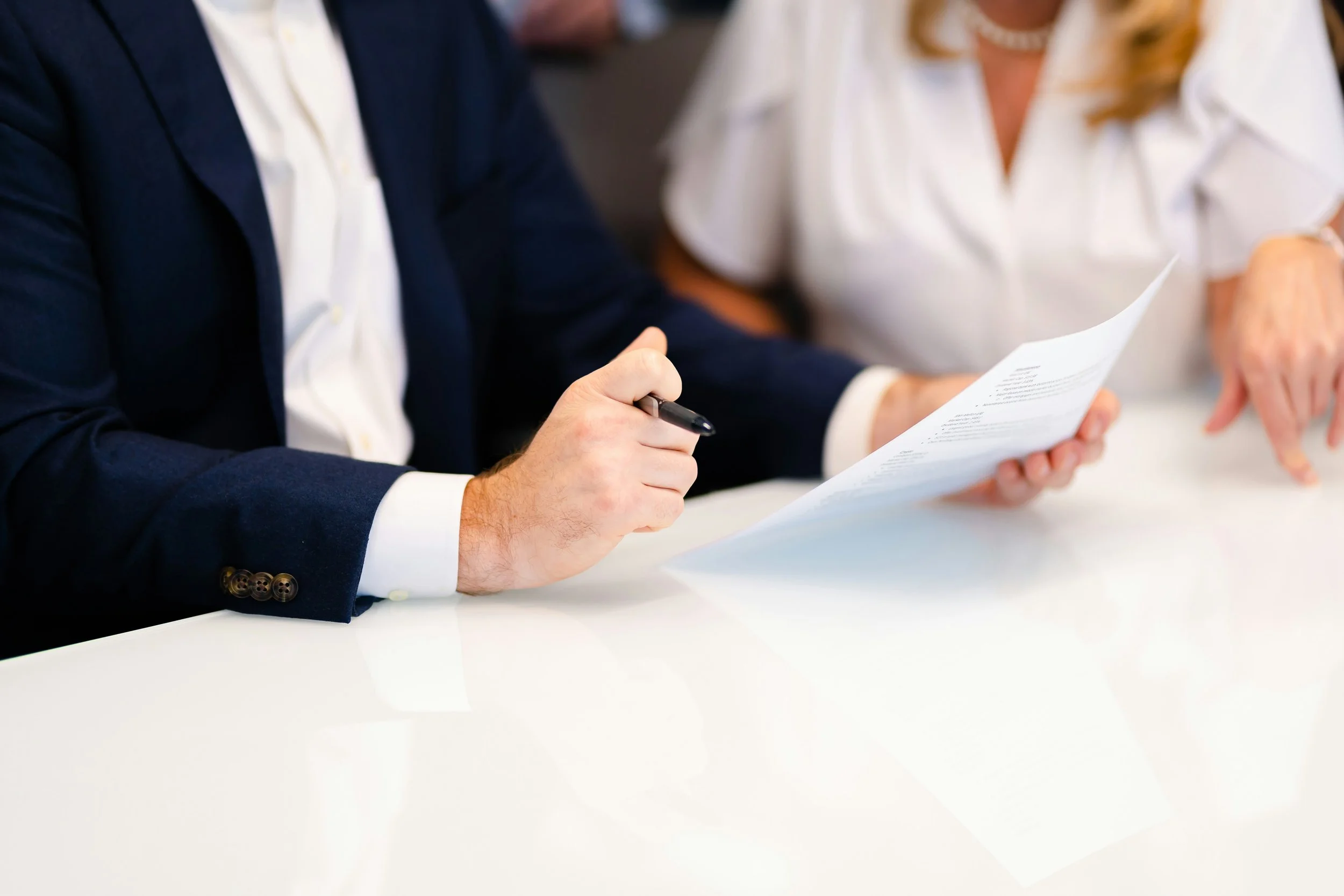 Two individuals reviewing a document together at a white table, one holding a pen and the other pointing at the paper.