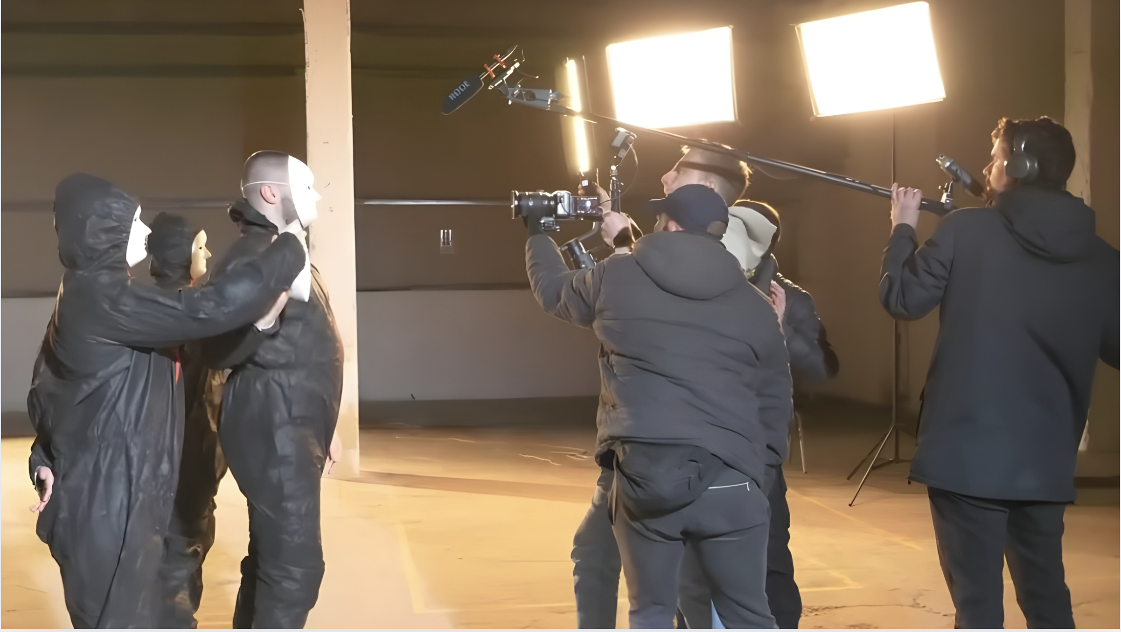 Film crew recording a scene with actors in black hoodies with masks, set in an indoor space with large bright lighting.