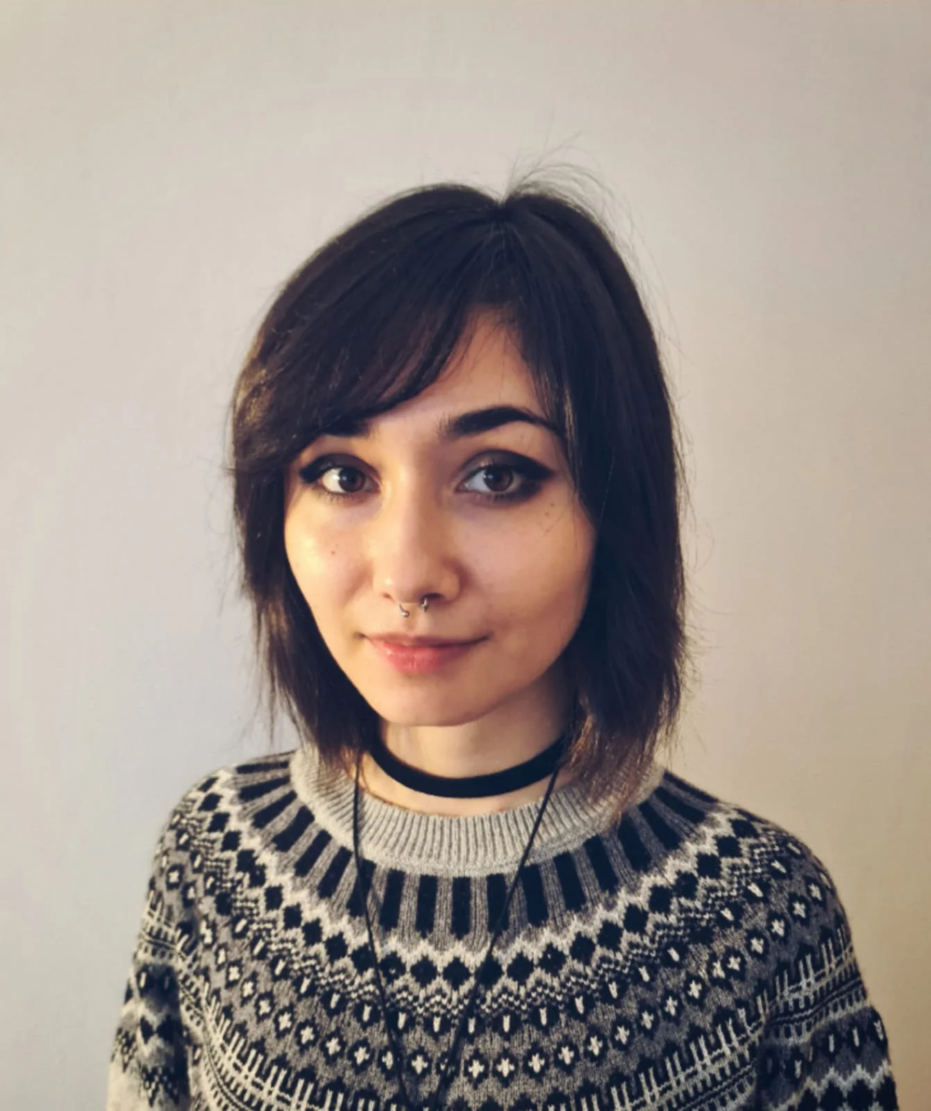 Young woman with dark hair wearing a patterned sweater, black choker, and headphones against a plain background.