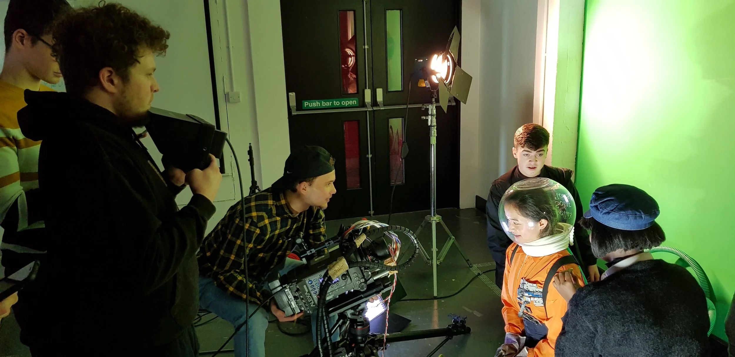 Group of people filming a scene with a green screen in a studio, one person operating a camera, others observing.