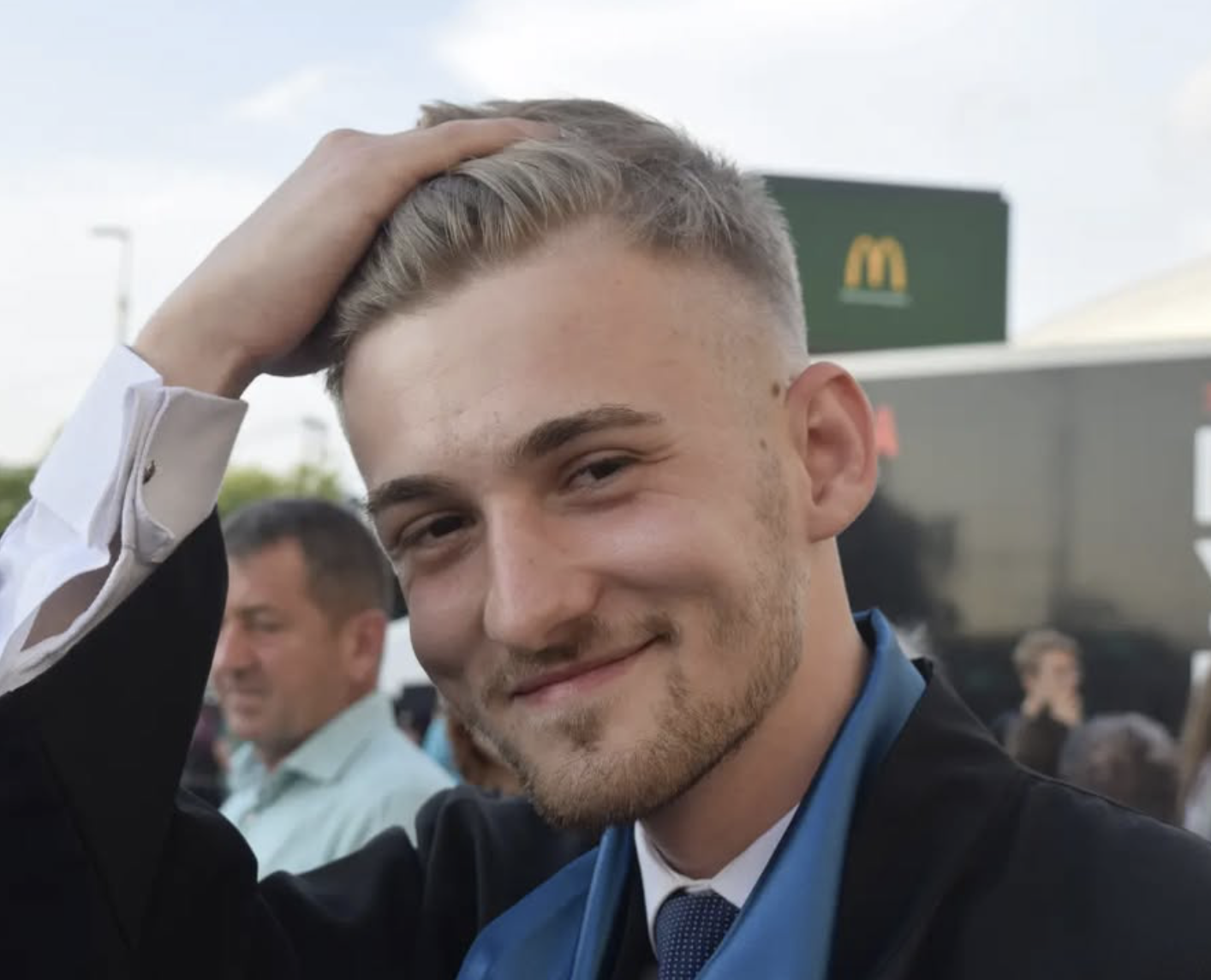 A young man with blond hair and a goatee smiling, touching his hair, wearing a black suit and blue dress shirt, in an outdoor setting with a McDonald's sign in the background.
