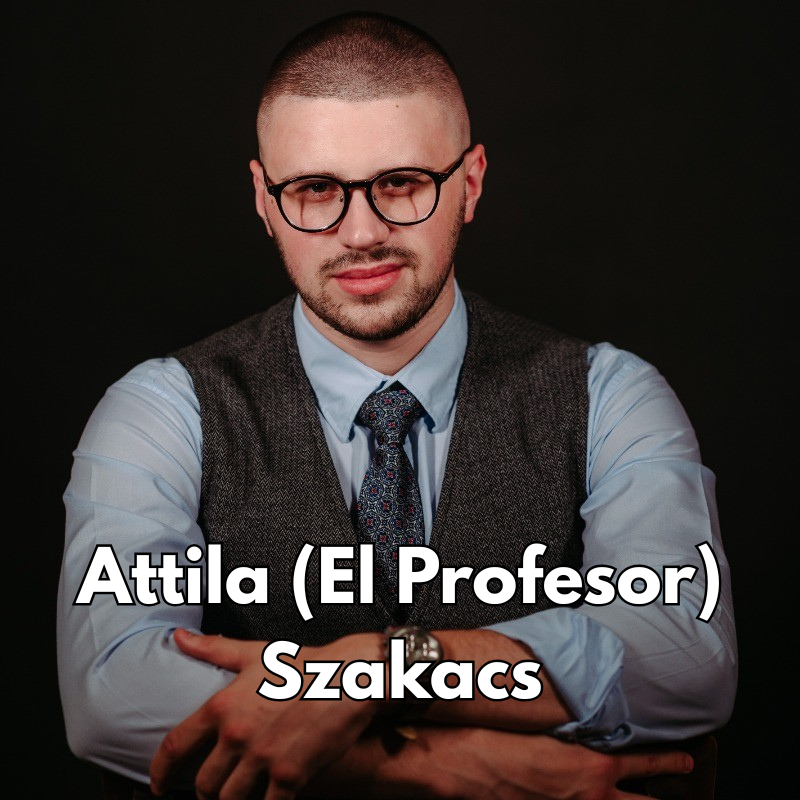 A young man with a beard and glasses, wearing a light blue shirt, a dark vest, and a patterned tie, sitting with folded arms against a black background, looking at the camera with a serious expression.