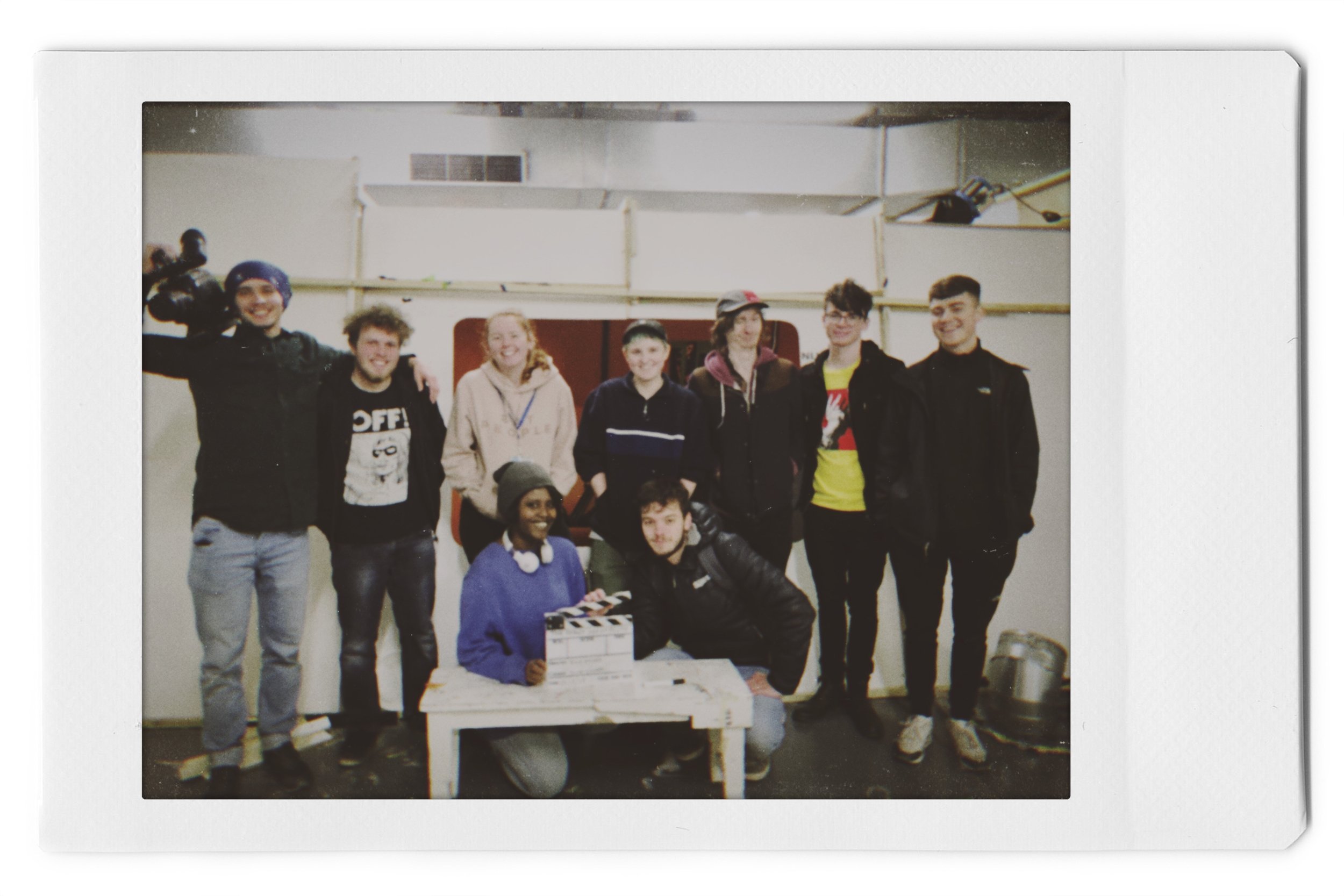 Group of nine young people posing for a photo in an indoor setting, with a white wall behind them and some equipment in the background. Two people are kneeling in front, and the rest are standing.