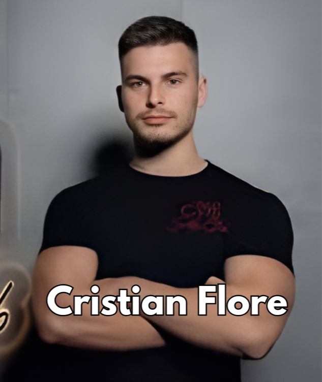 A young man with short dark hair and a slight beard, wearing a black T-shirt with red writing, standing with arms crossed against a plain gray background.