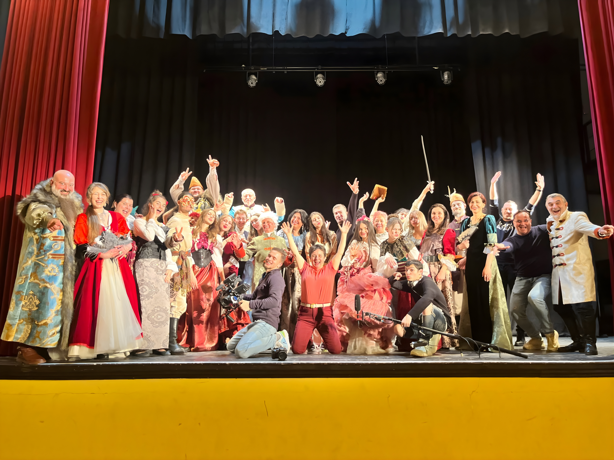 A large group of people in colorful costumes on a stage, celebrating and smiling, with some raising their hands and a few holding props like a sword and a book.