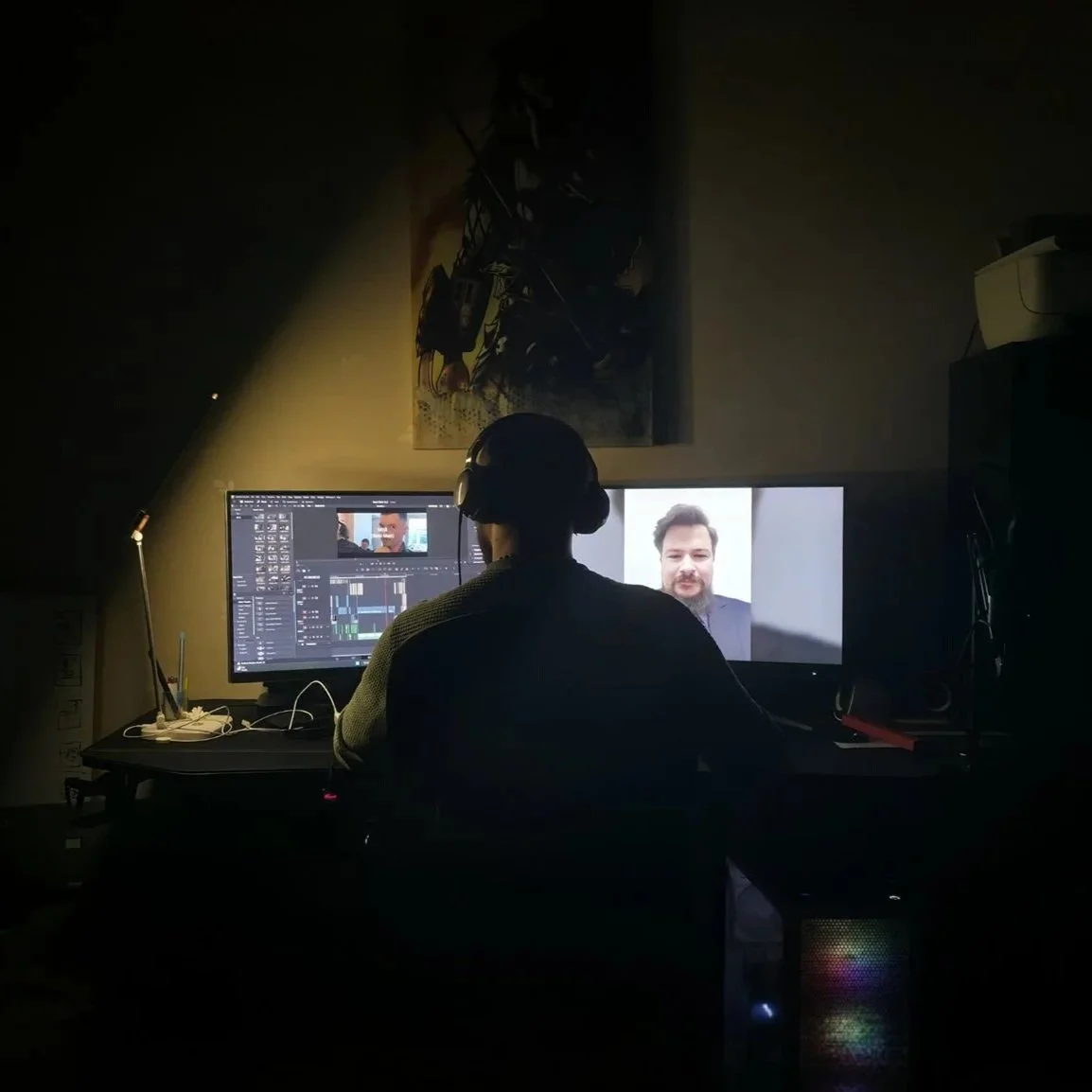 Person working on a video editing project at a desk in a dimly lit room, with dual monitors displaying video editing software and a man's face.